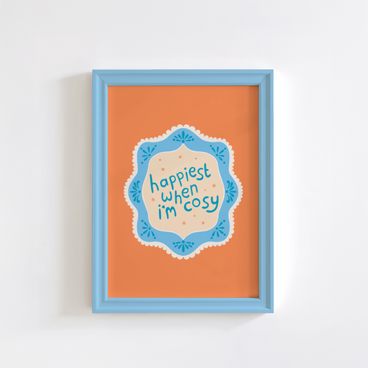 An orange print with a quote inside an illustrated blue decorative border - which says 'happiest when I'm cosy'.