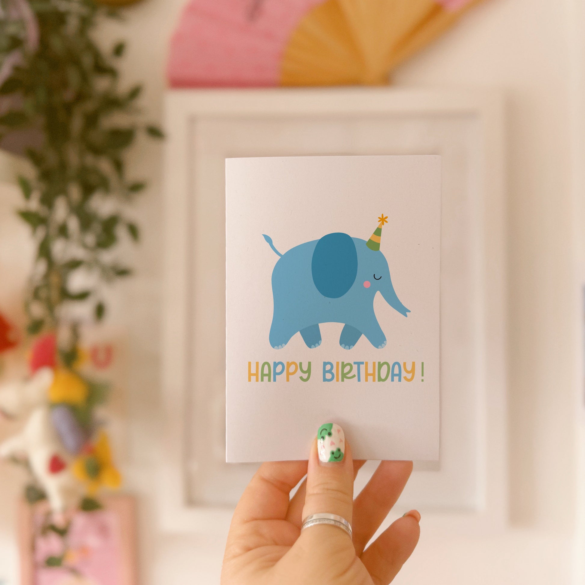 A hand holding a birthday card featuring a blue illustrated elephant with a party hat, and the text 'HAPPY BIRTHDAY!' on the front. The card is displayed against a background with various colours and patterns.