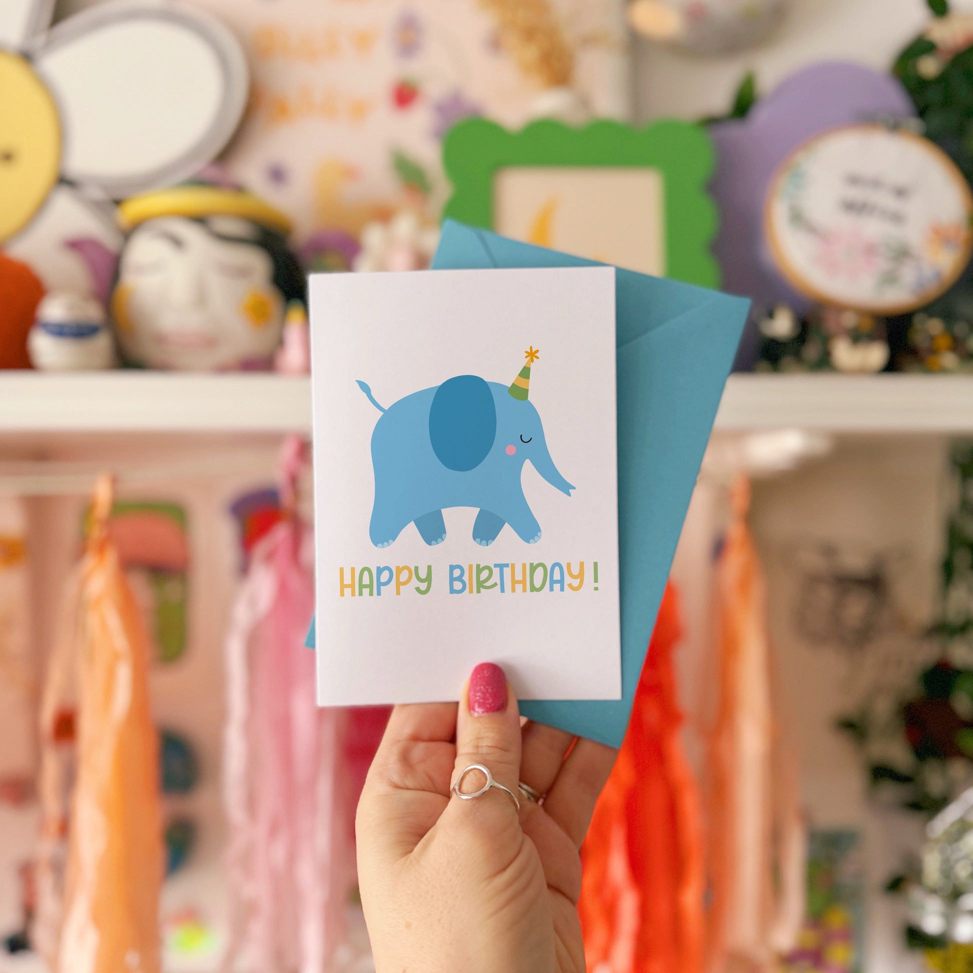 A hand holding a birthday card featuring a blue illustrated elephant with a party hat, and the text 'HAPPY BIRTHDAY!' on the front. The card is displayed against a background with various colours and patterns.