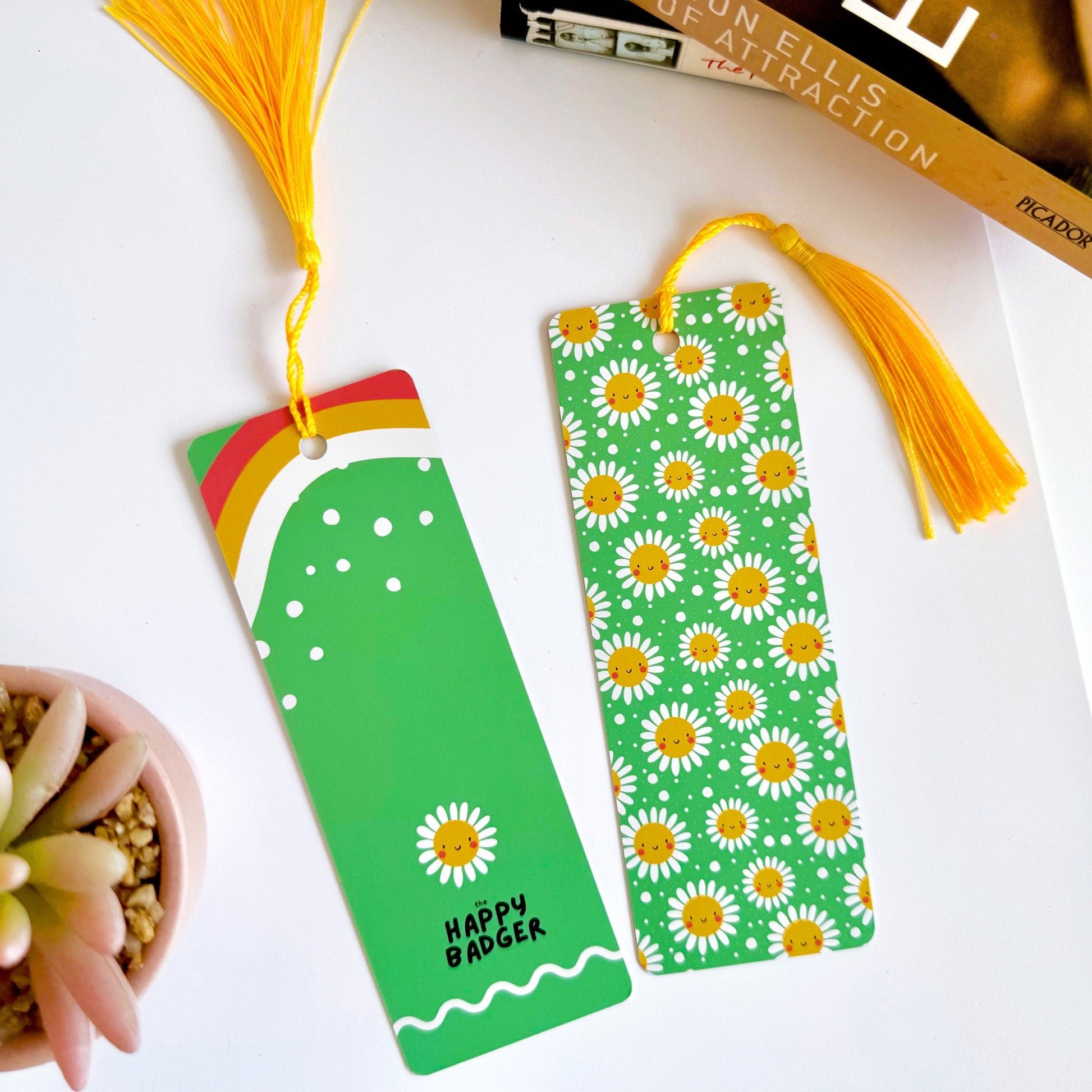 A picture of a green bookmark, with a coordinating yellow tassel, with a repeating pattern of white and yellow smiling flowers on it. Also shown is the rear side, which is solid green with pops of yellow and red, and the brand logo at the bottom.