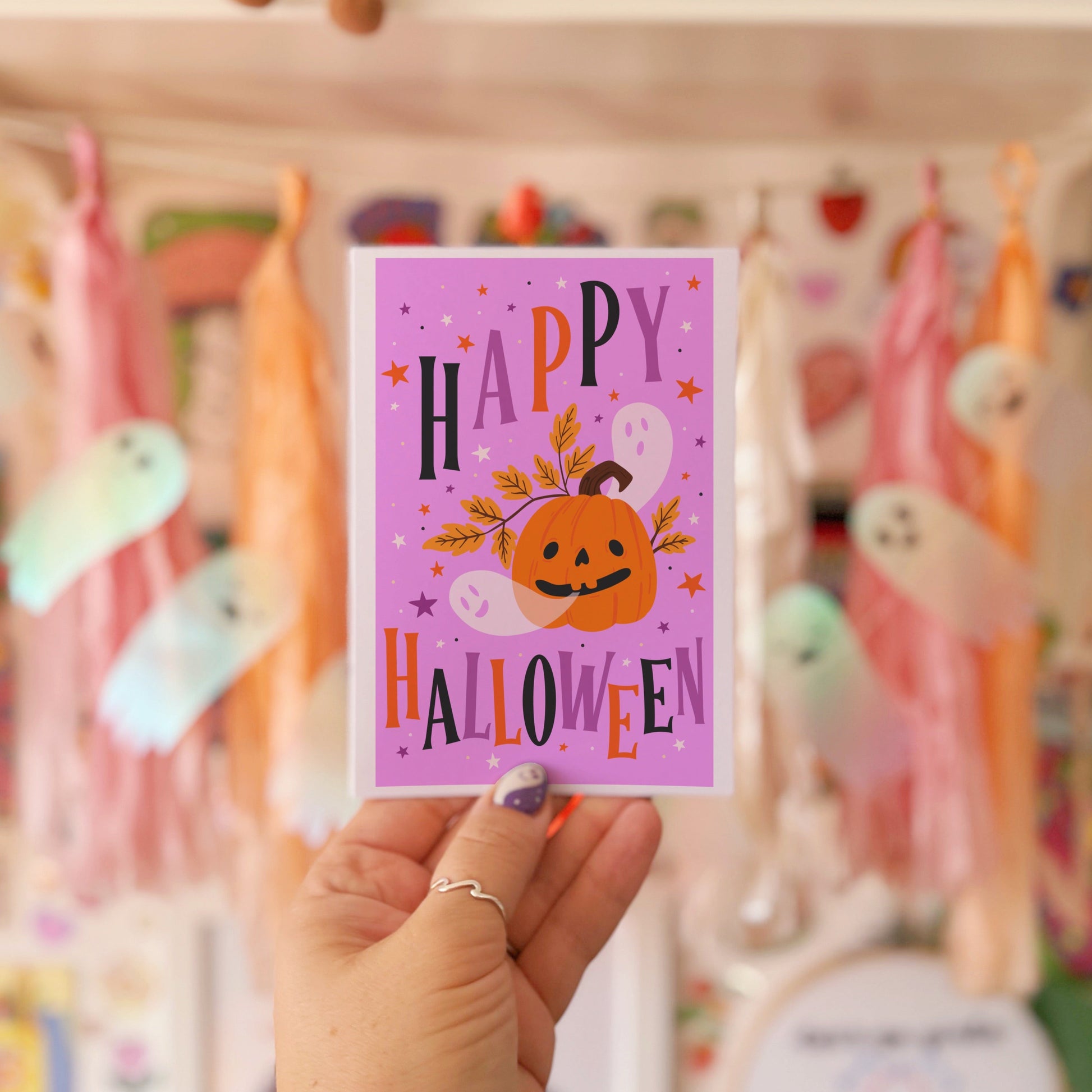 Hand holding a 'Happy Halloween' card in front of a colorful shelf with decorations.