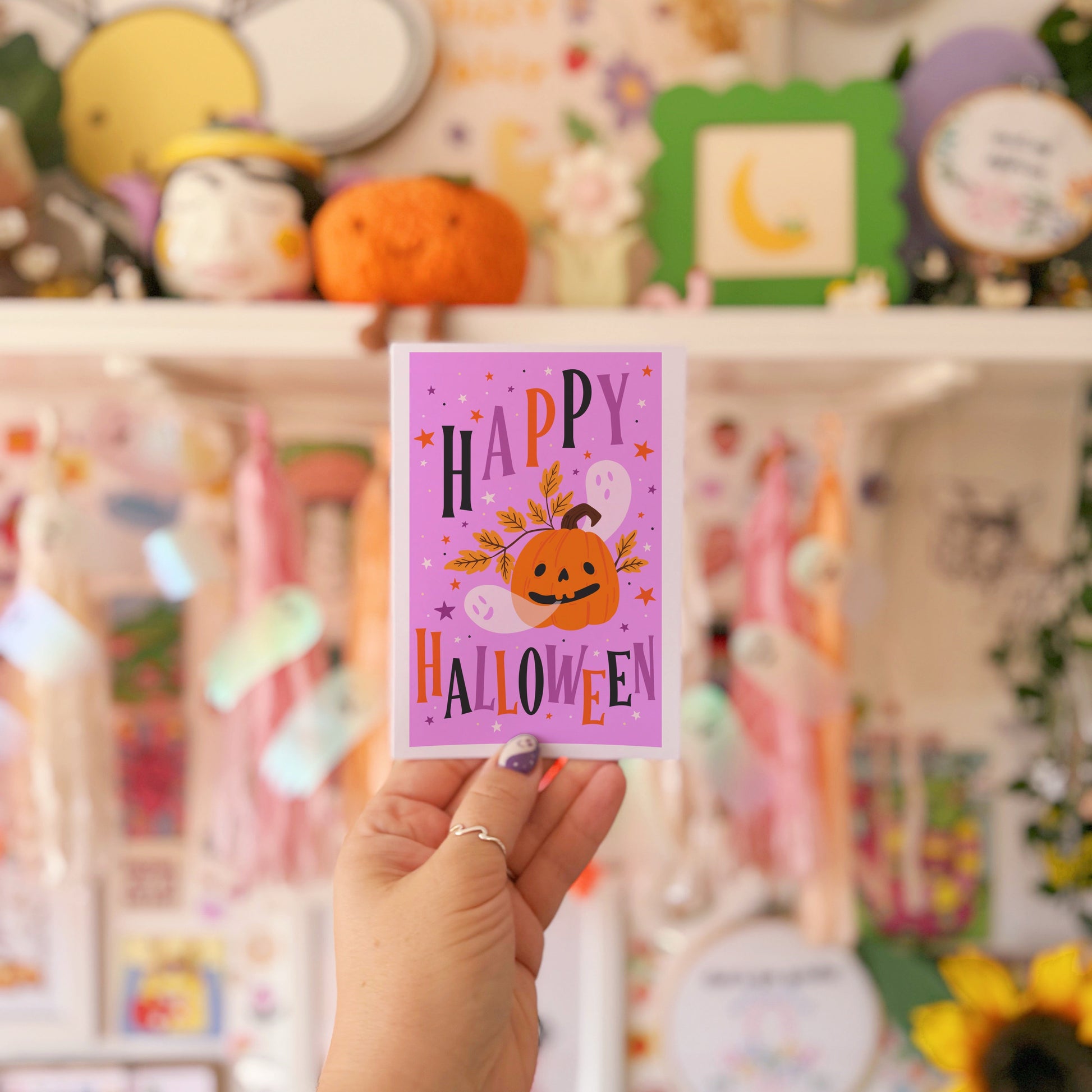 Hand holding a 'Happy Halloween' card in front of a decorative shelf with pumpkins and other items.
