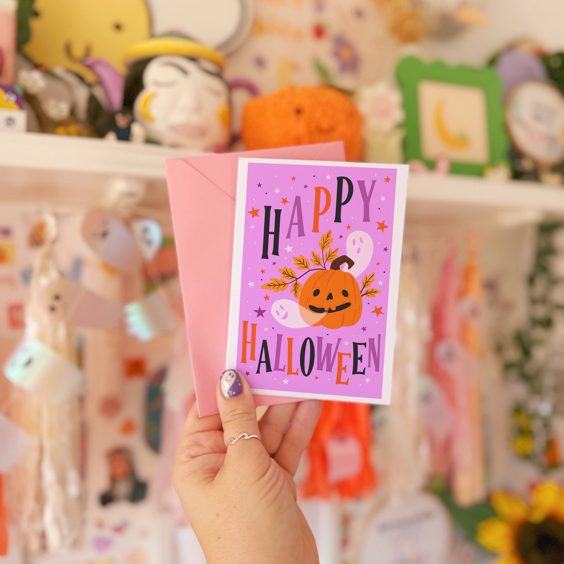 Hand holding a 'Happy Halloween' card with pumpkins in front of a colorful background with toys and decorations.