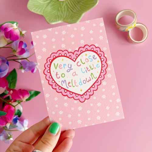 Hand holding a pink card with a heart design and text on a pink background with flowers and tape.
