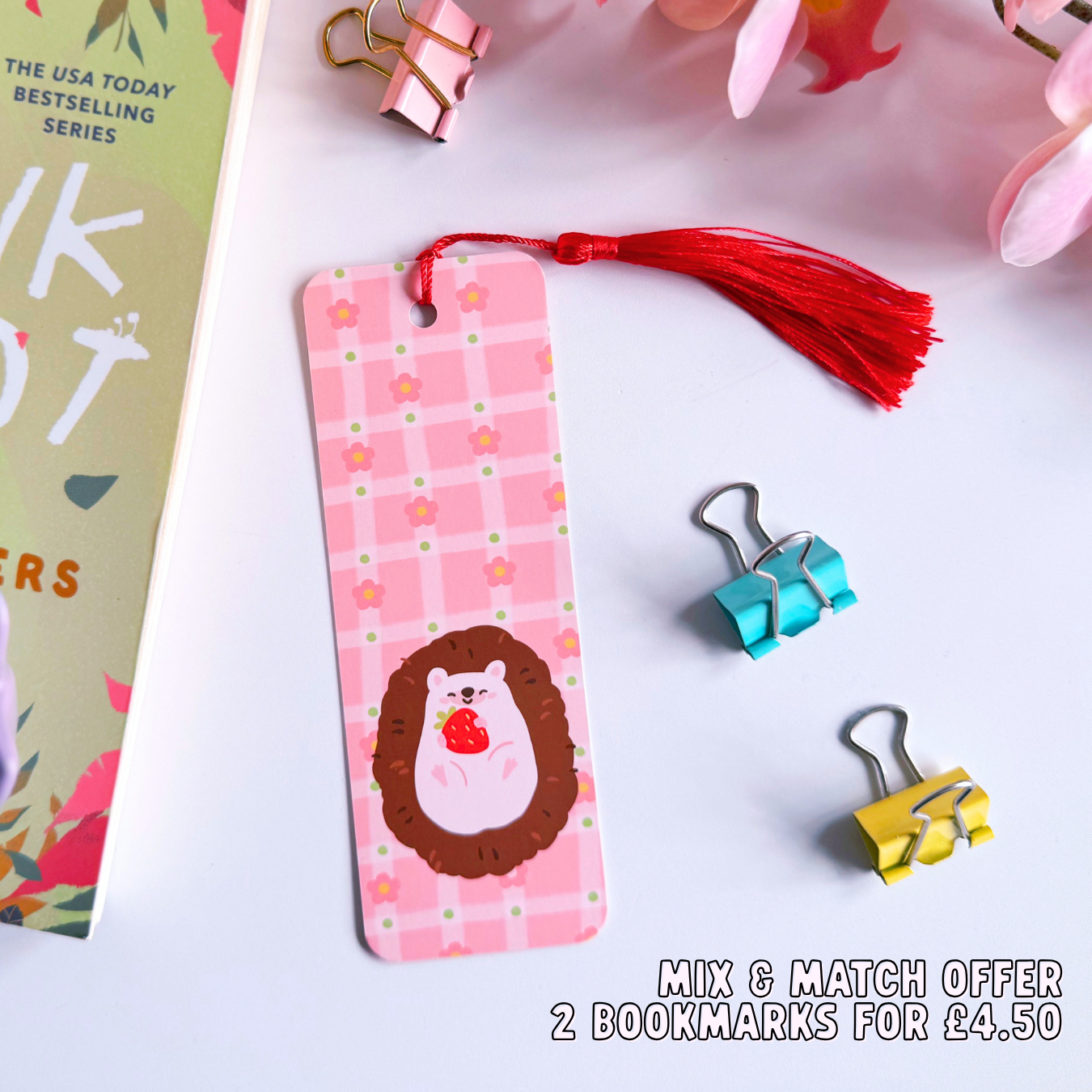 A pink bookmark with a red tassel, with little pink daisies dotted around a little hedgehog holding a strawberry. The back of the bookmark has the same pattern, but an illustration of daisies and the branding logo.