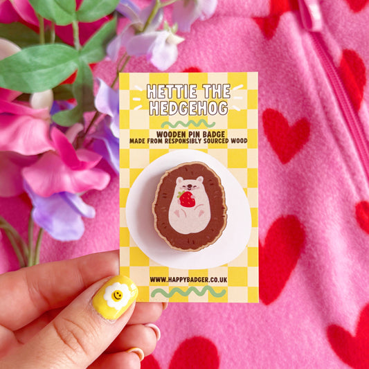A wooden pin badge featuring a cute hedgehog curled up in a ball, with a little smiling face, holding a red strawberry in her arms.