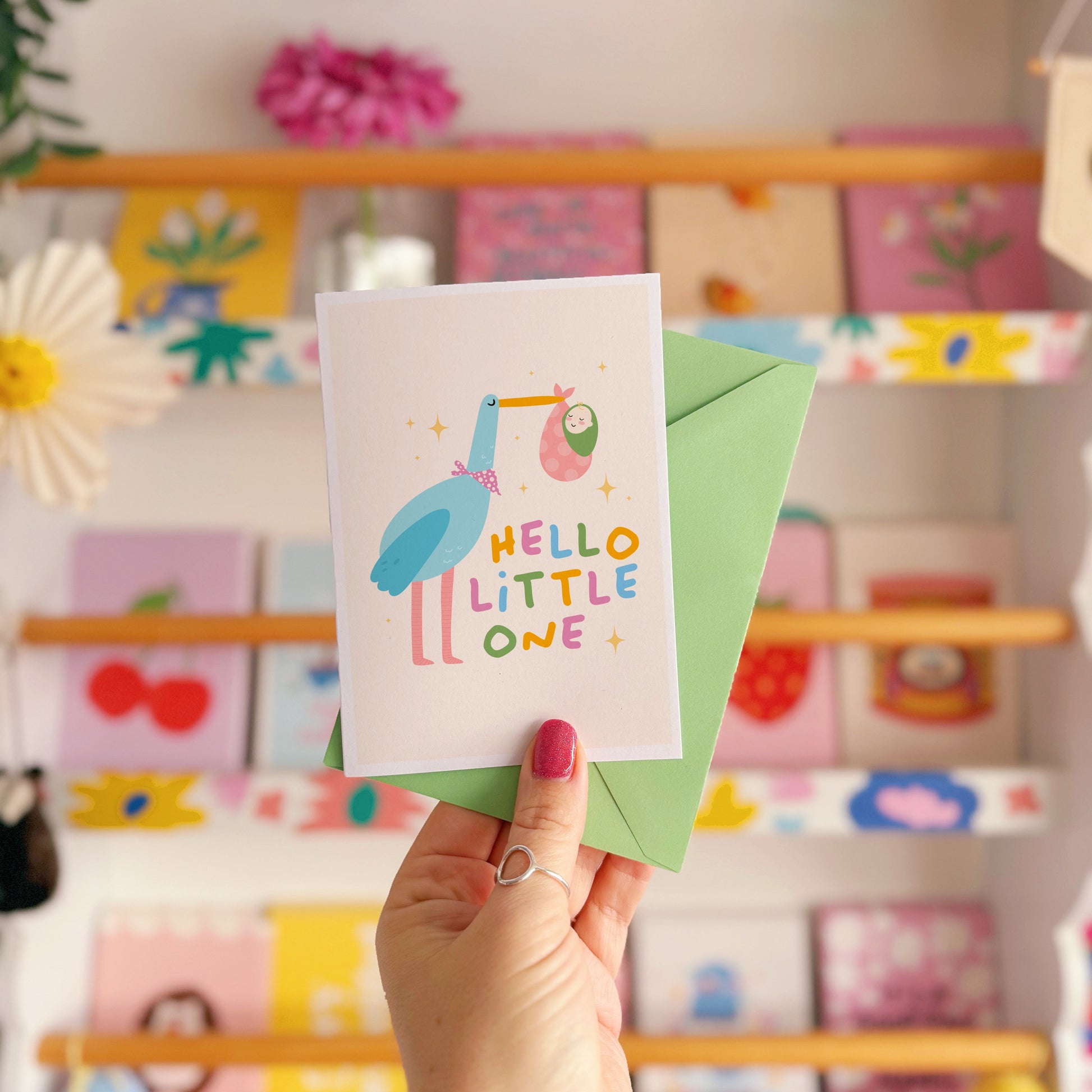 A greetings card with an illustration of a stalk carrying a baby, with the words 'hello little one' in colourful tones.