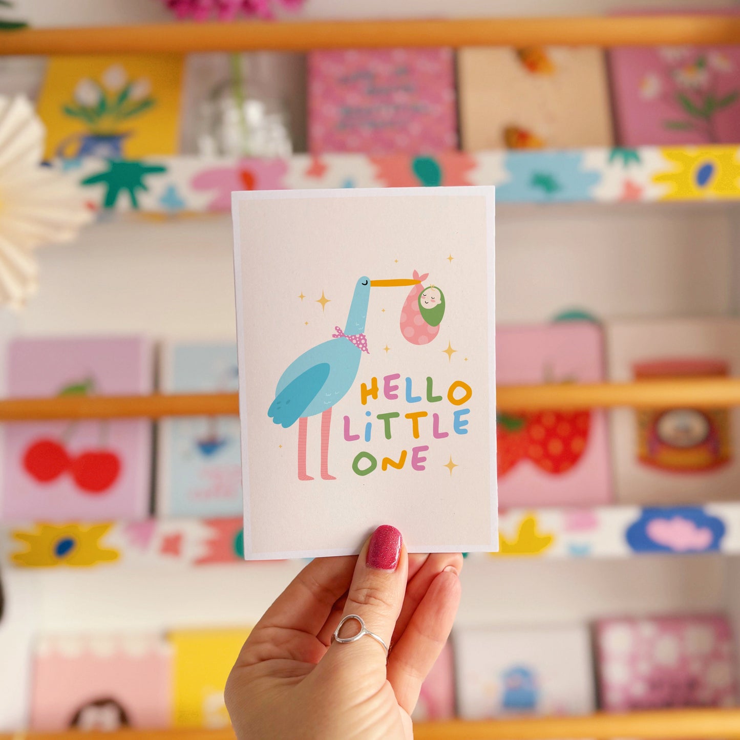 A greetings card with an illustration of a stalk carrying a baby, with the words 'hello little one' in colourful tones.