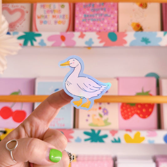 A sticker with an illustration of a white goose, smiling. The sticker has a blue border and the backing has an easy peel lip.