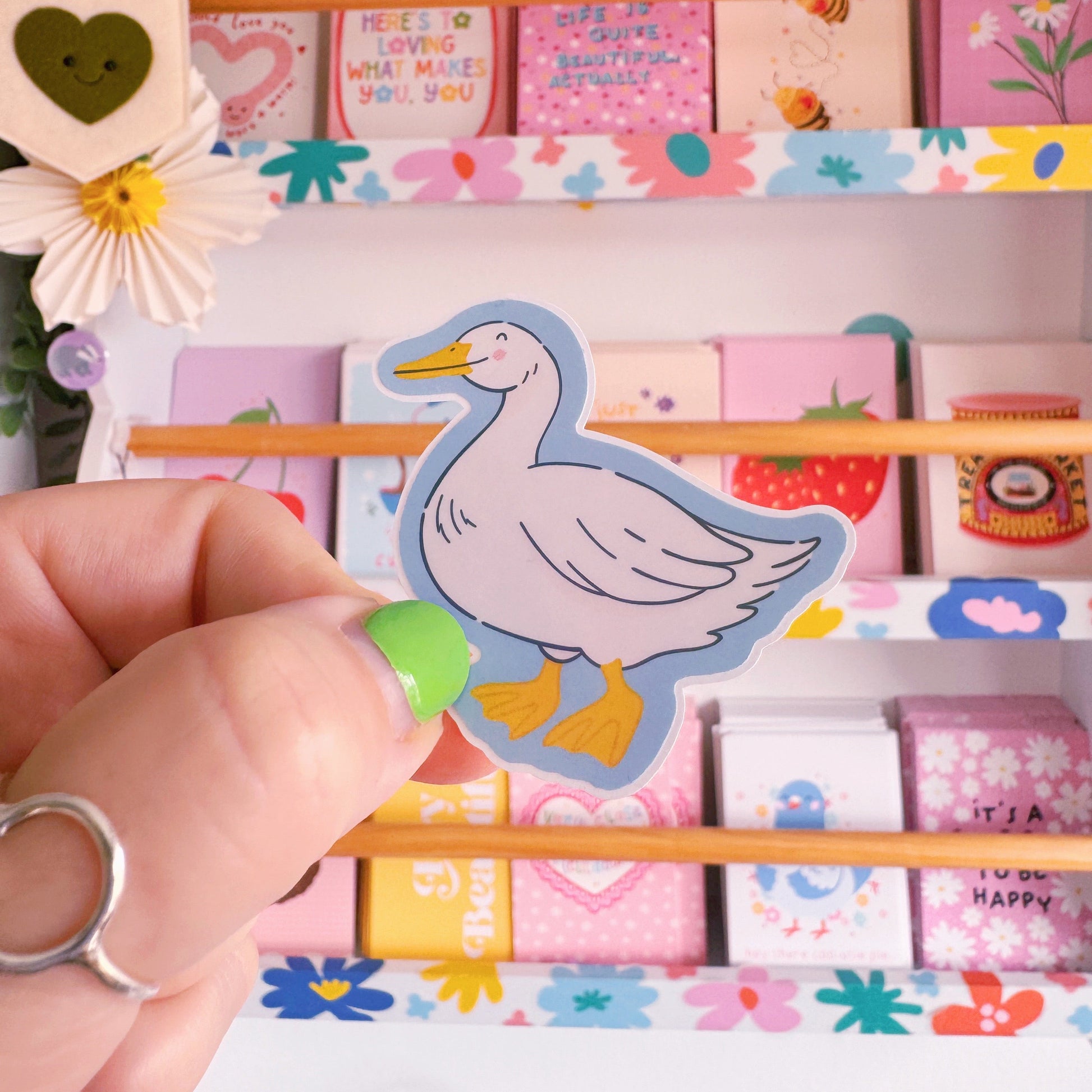 A sticker with an illustration of a white goose, smiling. The sticker has a blue border and the backing has an easy peel lip.