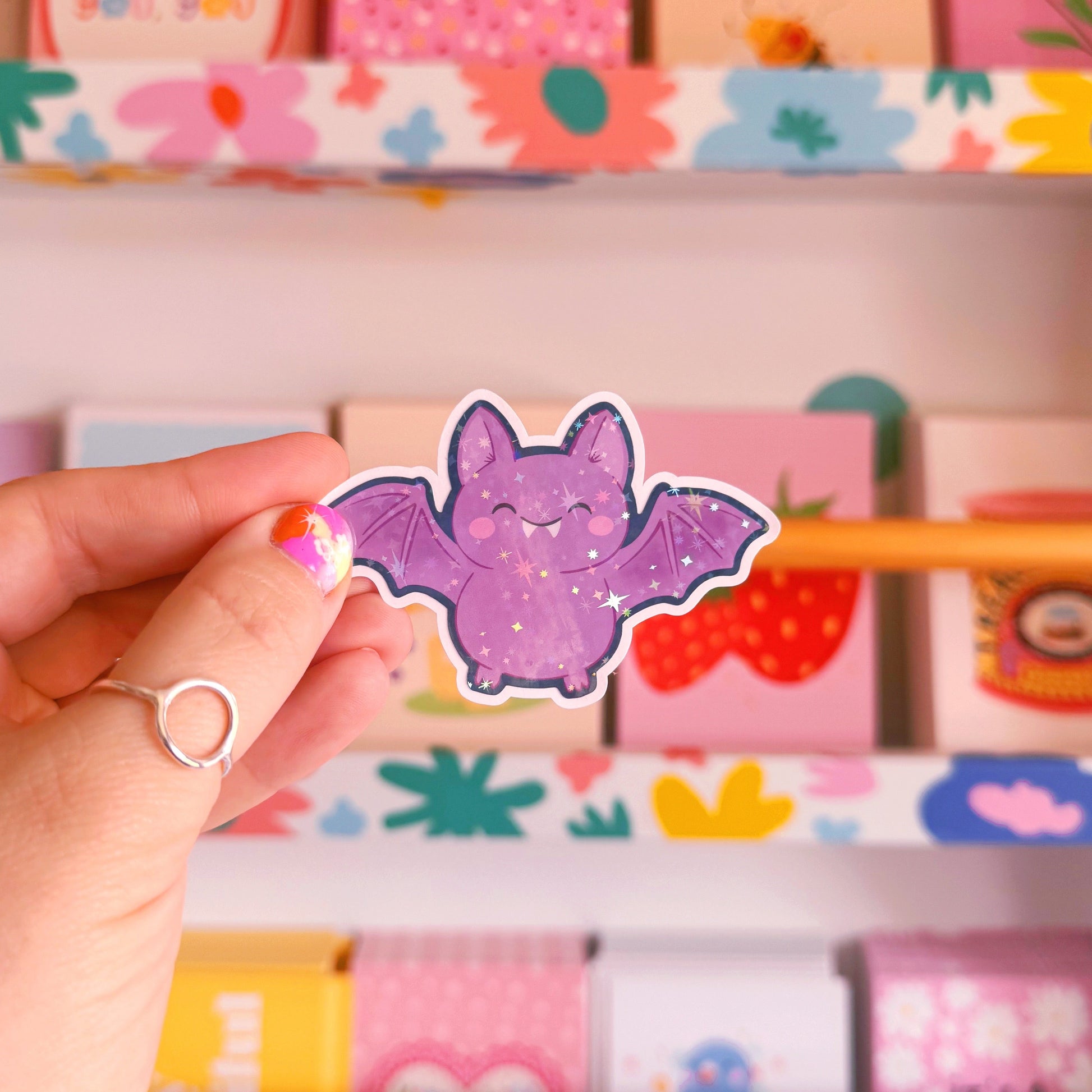 A holographic halloween themed sticker featuring an illustration of a purple bat with a navy border.