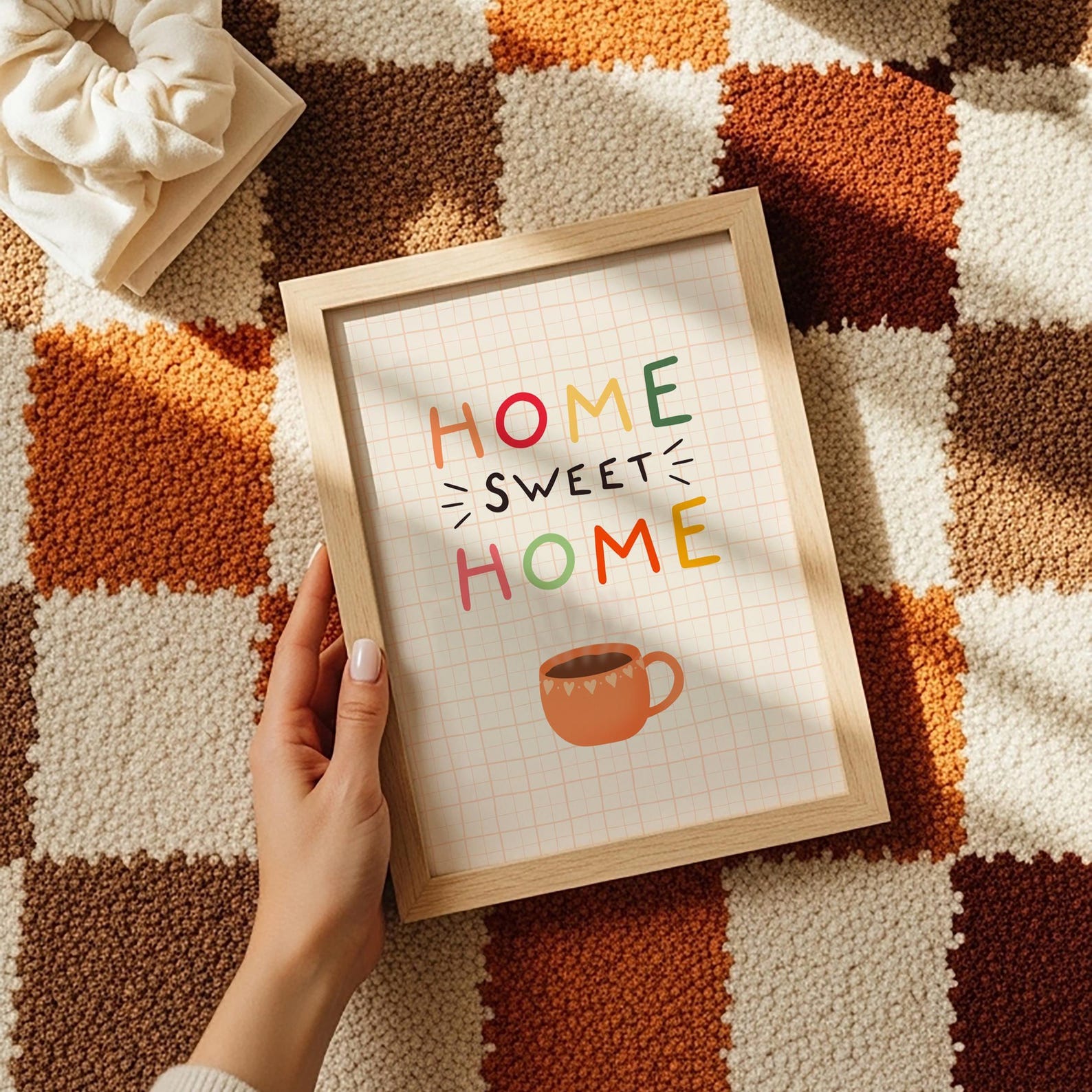 A cosy quote print that reads 'home sweet home' with an orange mug of coffee underneath. The background of the print is cream with a soft orange checkered pattern.