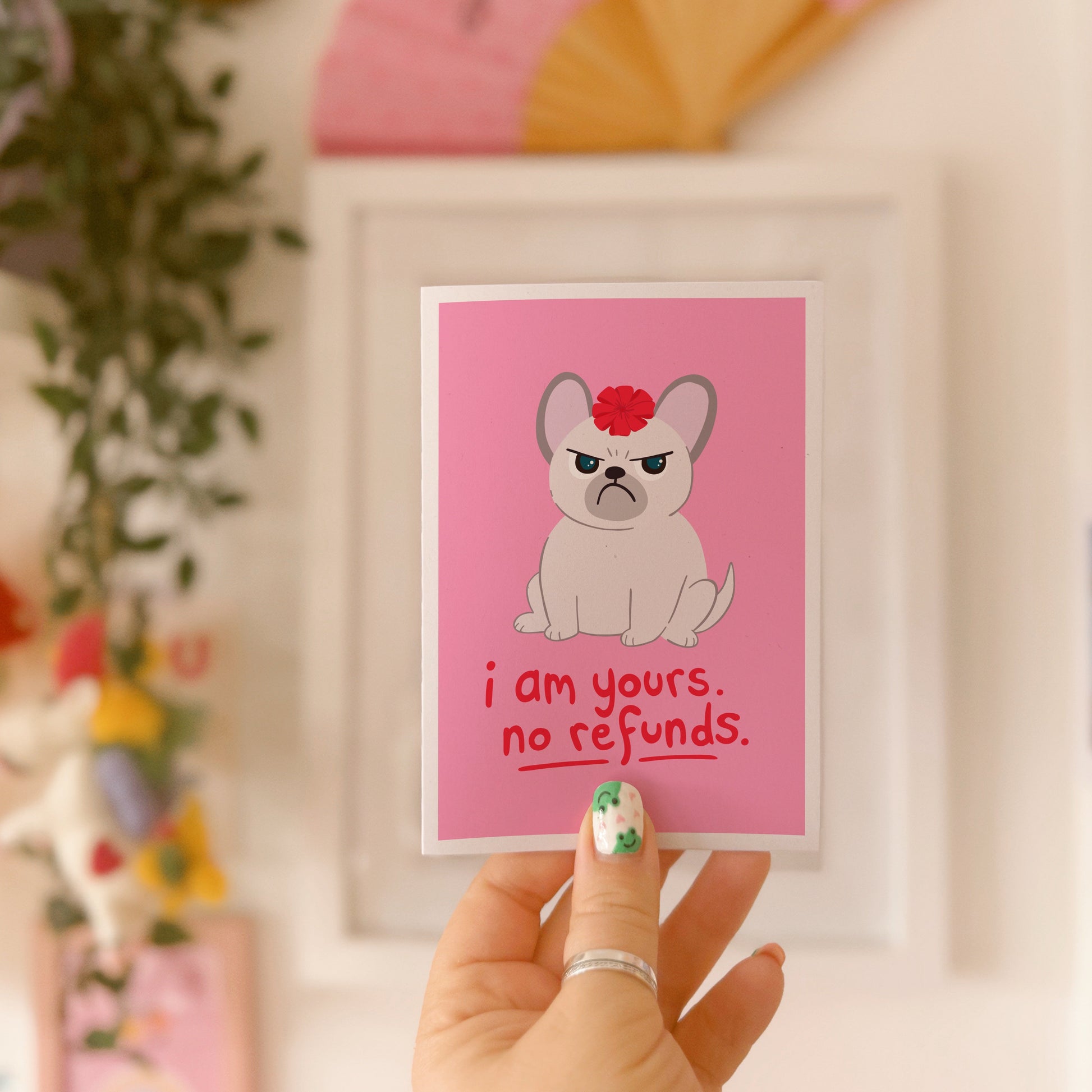 A pink greetings card with an illustration of a grumpy dog with a bow on it's head, and text underneath that reads 'I am yours, no refunds.'