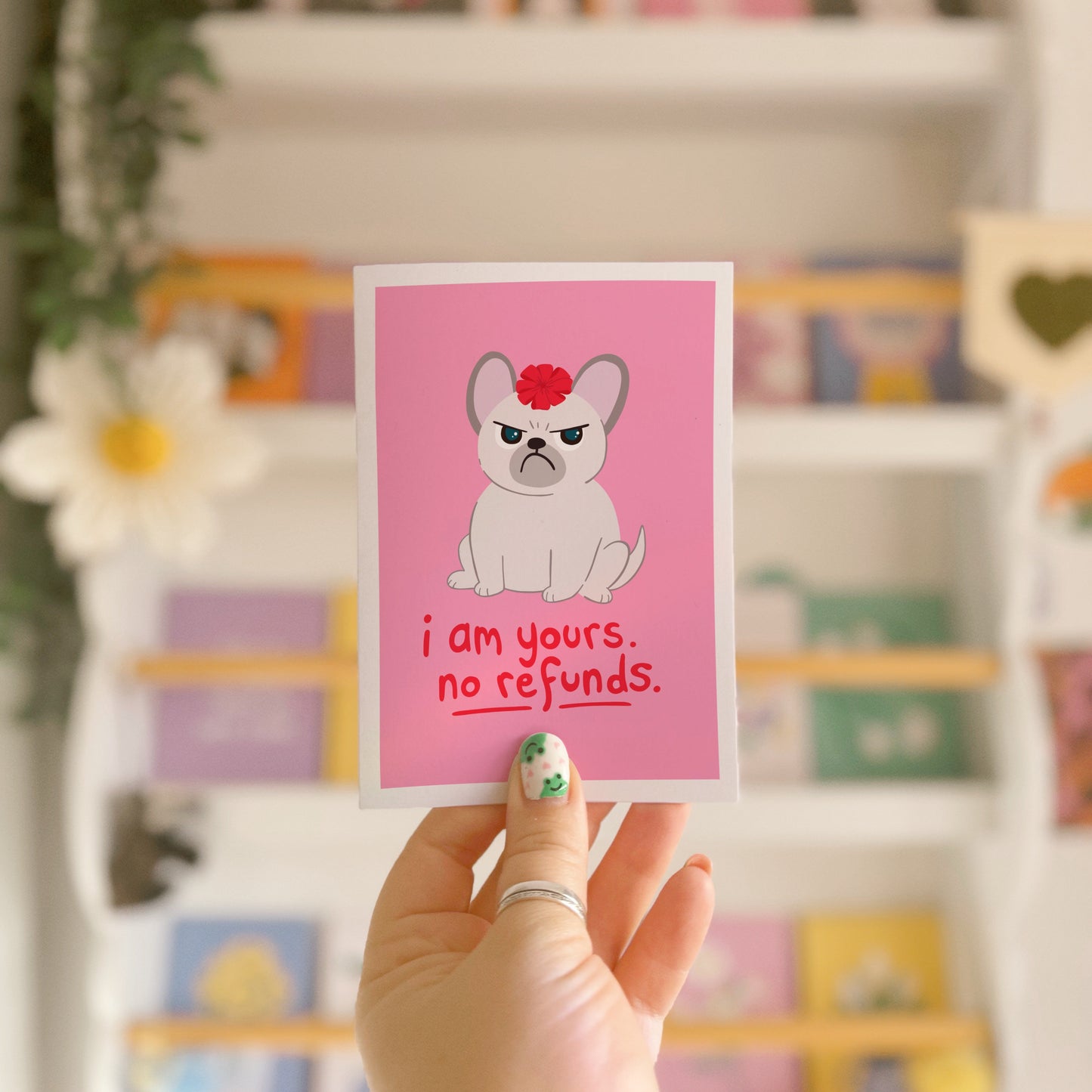 A pink greetings card with an illustration of a grumpy dog with a bow on it's head, and text underneath that reads 'I am yours, no refunds.'