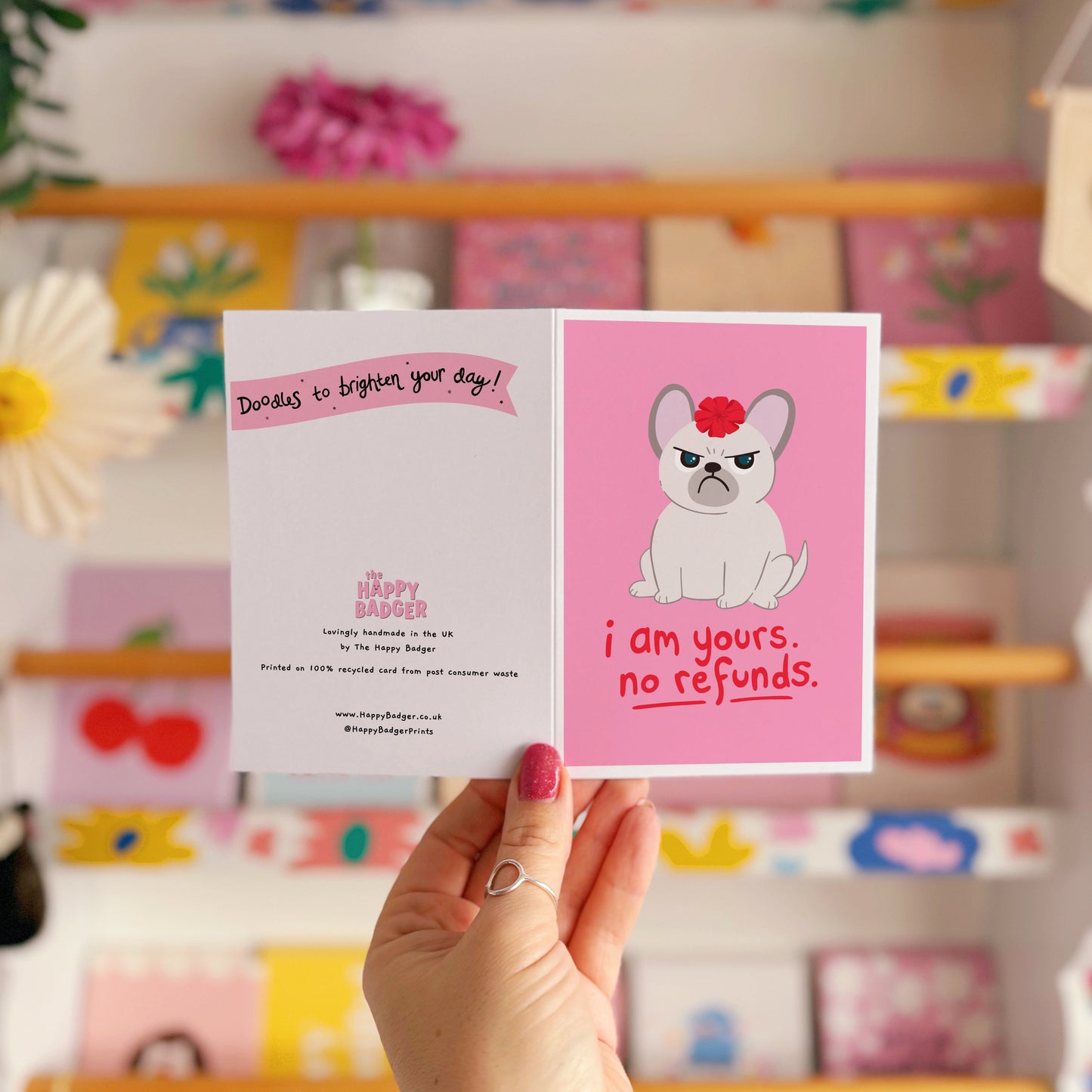 A pink greetings card with an illustration of a grumpy dog with a bow on it's head, and text underneath that reads 'I am yours, no refunds.'