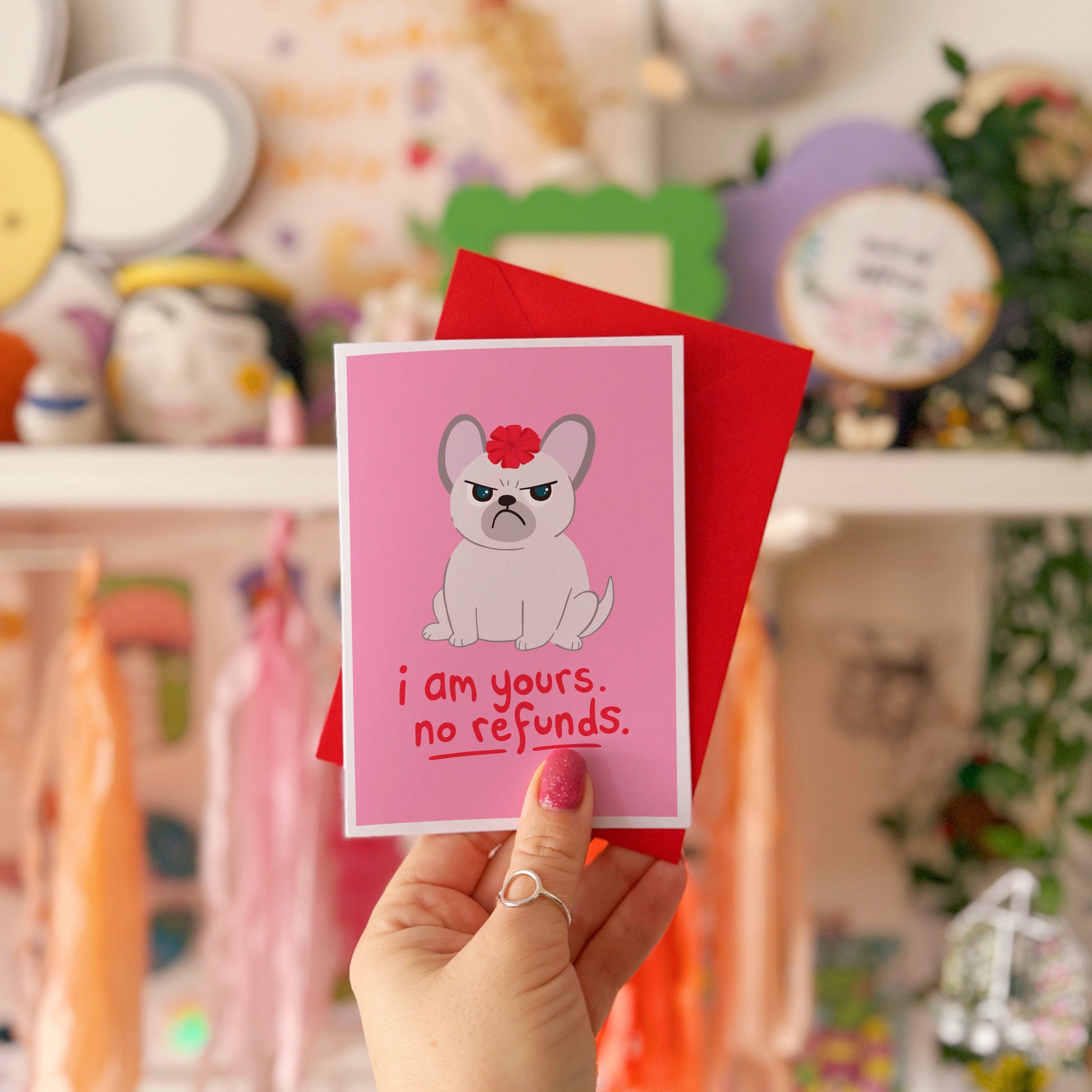 A pink greetings card with an illustration of a grumpy dog with a bow on it's head, and text underneath that reads 'I am yours, no refunds.'