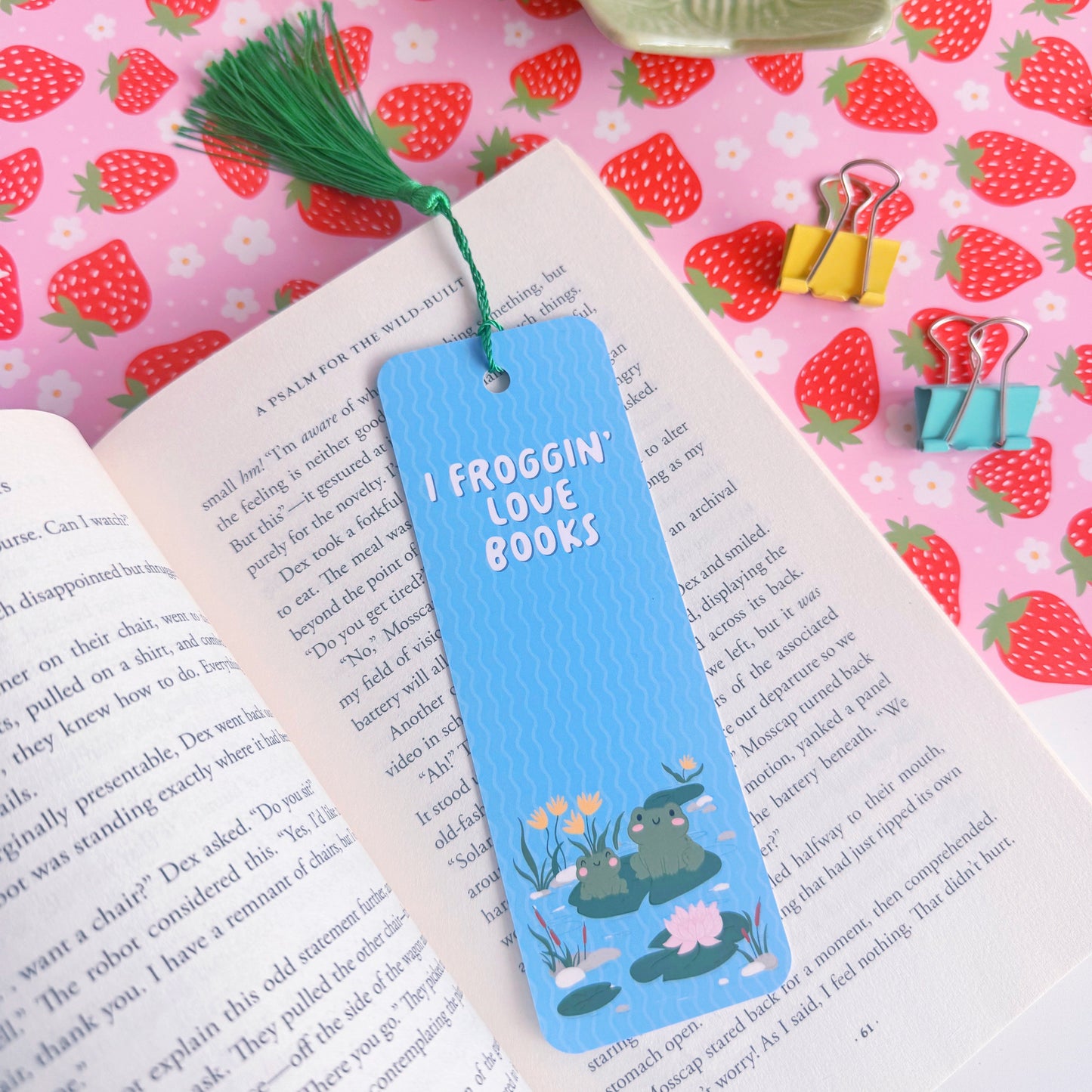 A blue bookmark with a cute frog illustration to both the front and back of the bookmark, on the front of the bookmark it says 'I Froggin' Love Books' and on the back of the bookmark is the brand logo.