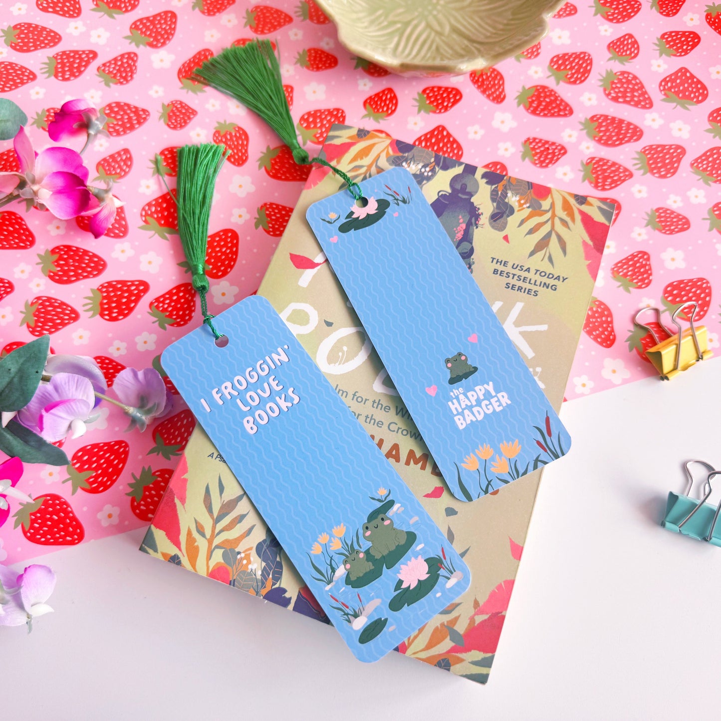 A blue bookmark with a cute frog illustration to both the front and back of the bookmark, on the front of the bookmark it says 'I Froggin' Love Books' and on the back of the bookmark is the brand logo.