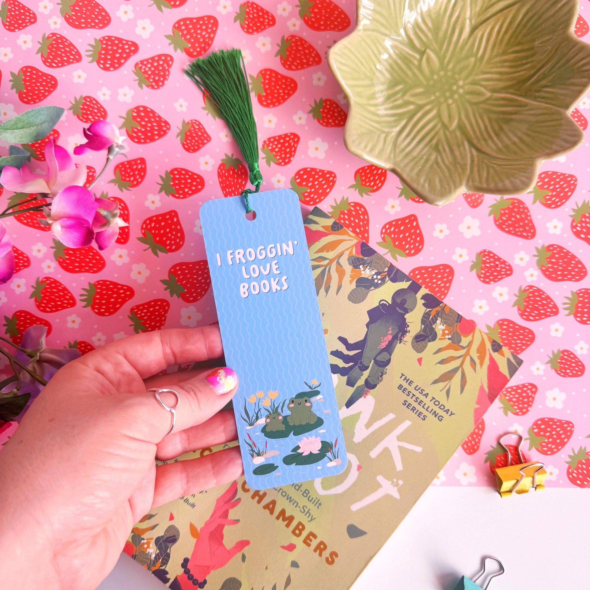 A blue bookmark with a cute frog illustration to both the front and back of the bookmark, on the front of the bookmark it says 'I Froggin' Love Books' and on the back of the bookmark is the brand logo.