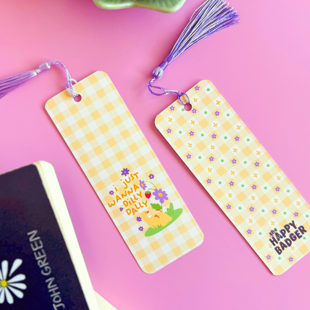 A yellow gingham bookmark with an illustration of a little duck with purple flowers. The text on the bookmark reads 'I just wanna dilly dally'.