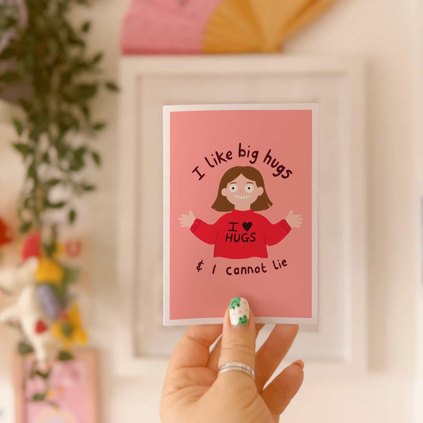 A pinkish red card with an illustration of a slightly creepy looking woman wearing a red jumper that says 'I love hugs' and text around her saying 'I like big hugs and I cannot lie'.