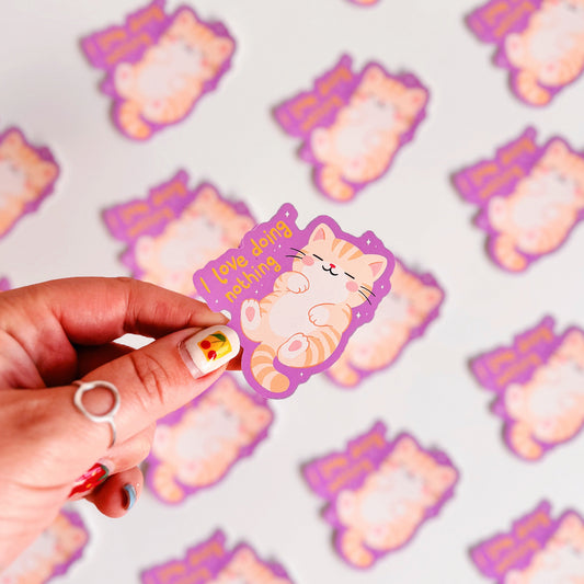Several purple vinyl stickers with an illustration of a sleeping cat, next to a hand lettered quote that reads 'I love doing nothing'.