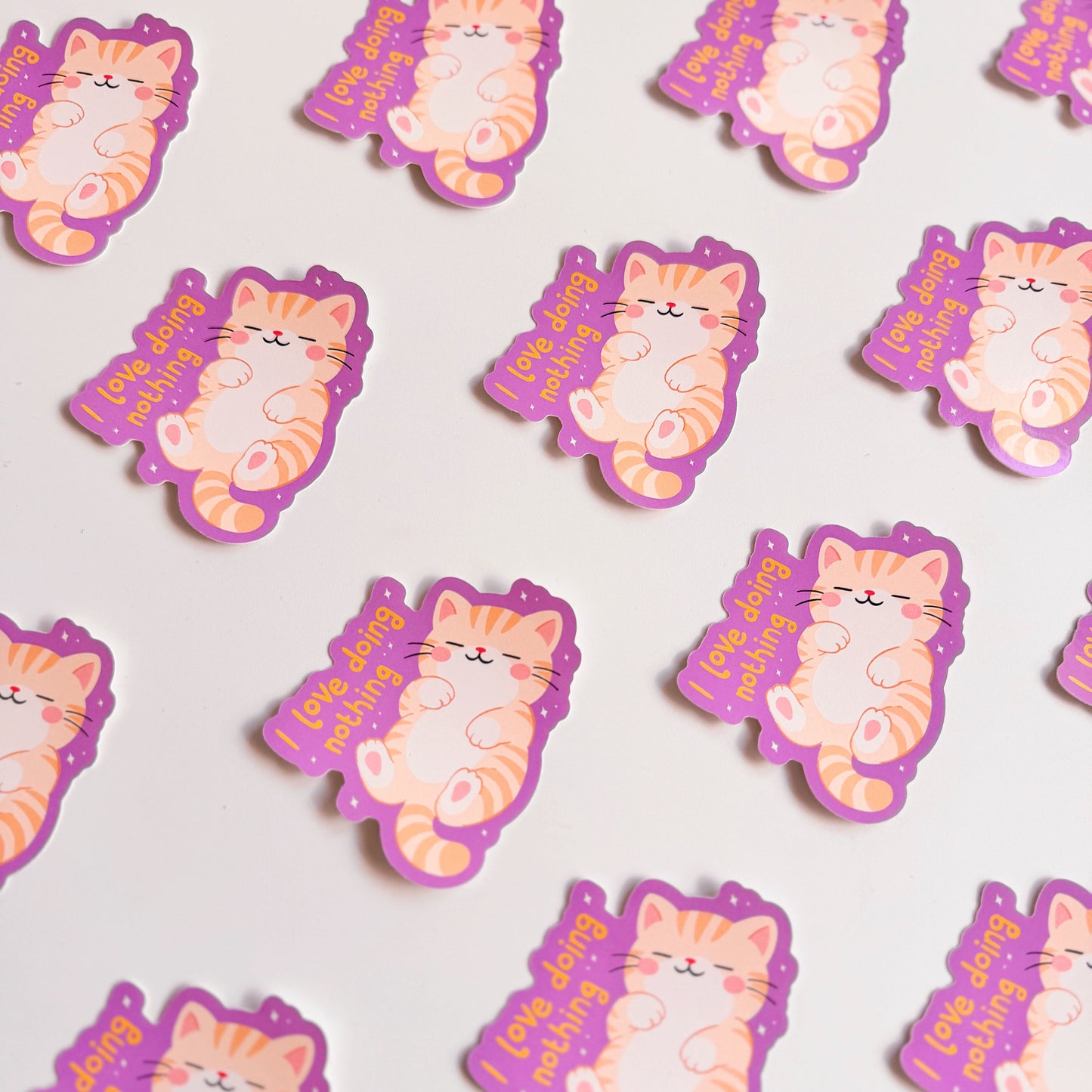 Several purple vinyl stickers with an illustration of a sleeping cat, next to a hand lettered quote that reads 'I love doing nothing'.