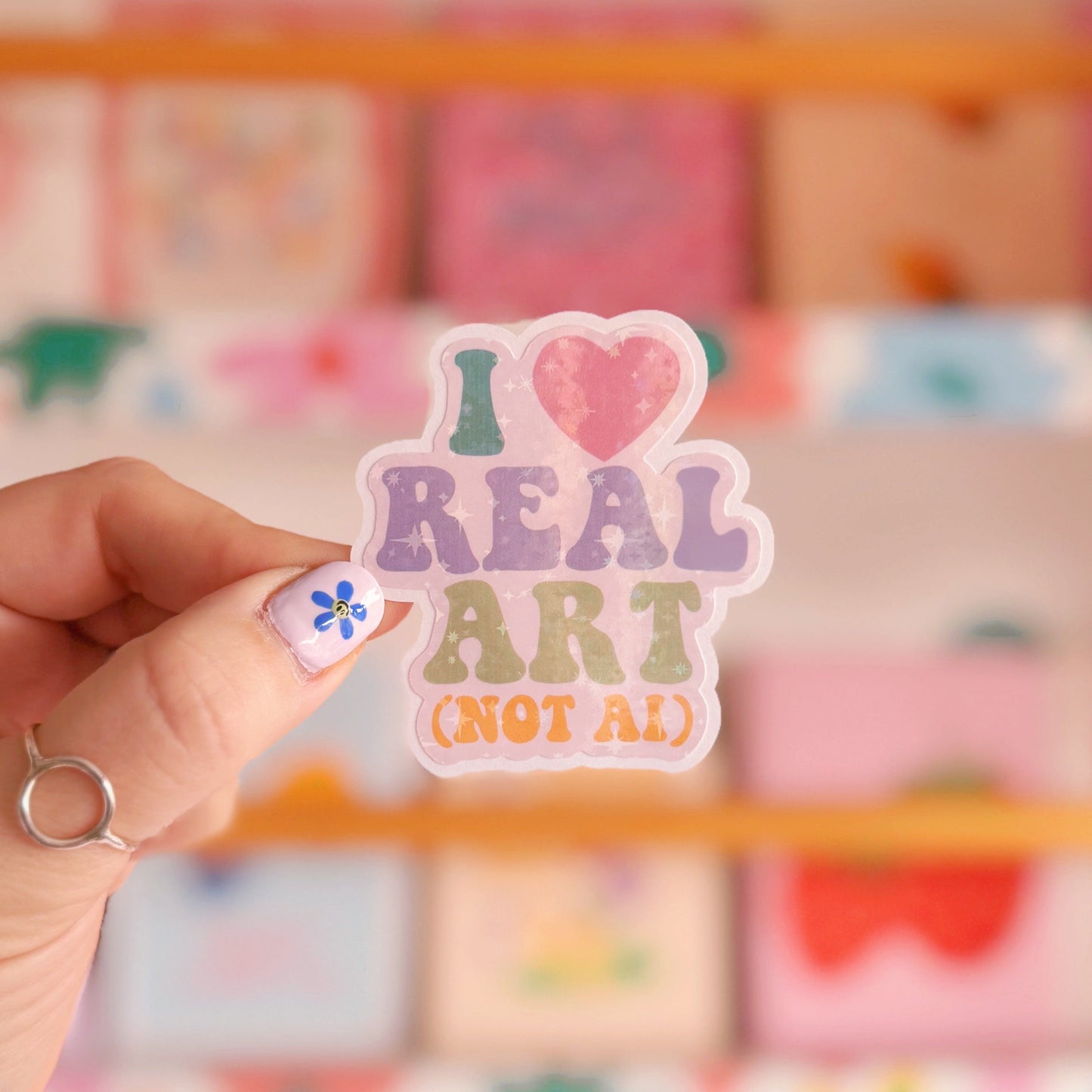 A holographic sticker that says 'I heart real art, (not AI)' in support of all artists struggling with the rise of generative AI art slop.