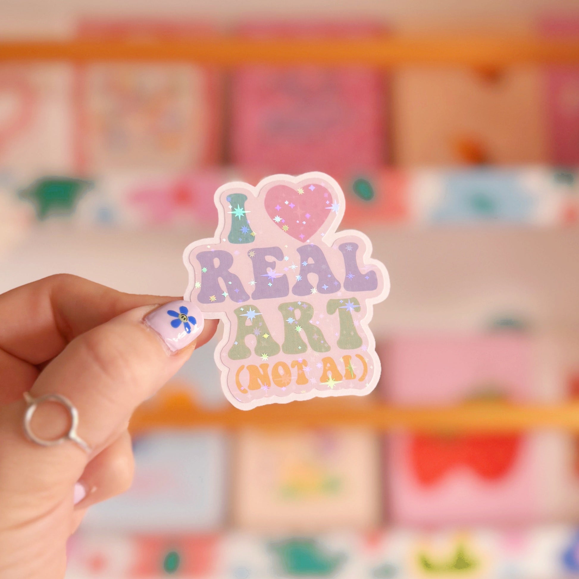 A holographic sticker that says 'I heart real art, (not AI)' in support of all artists struggling with the rise of generative AI art slop.