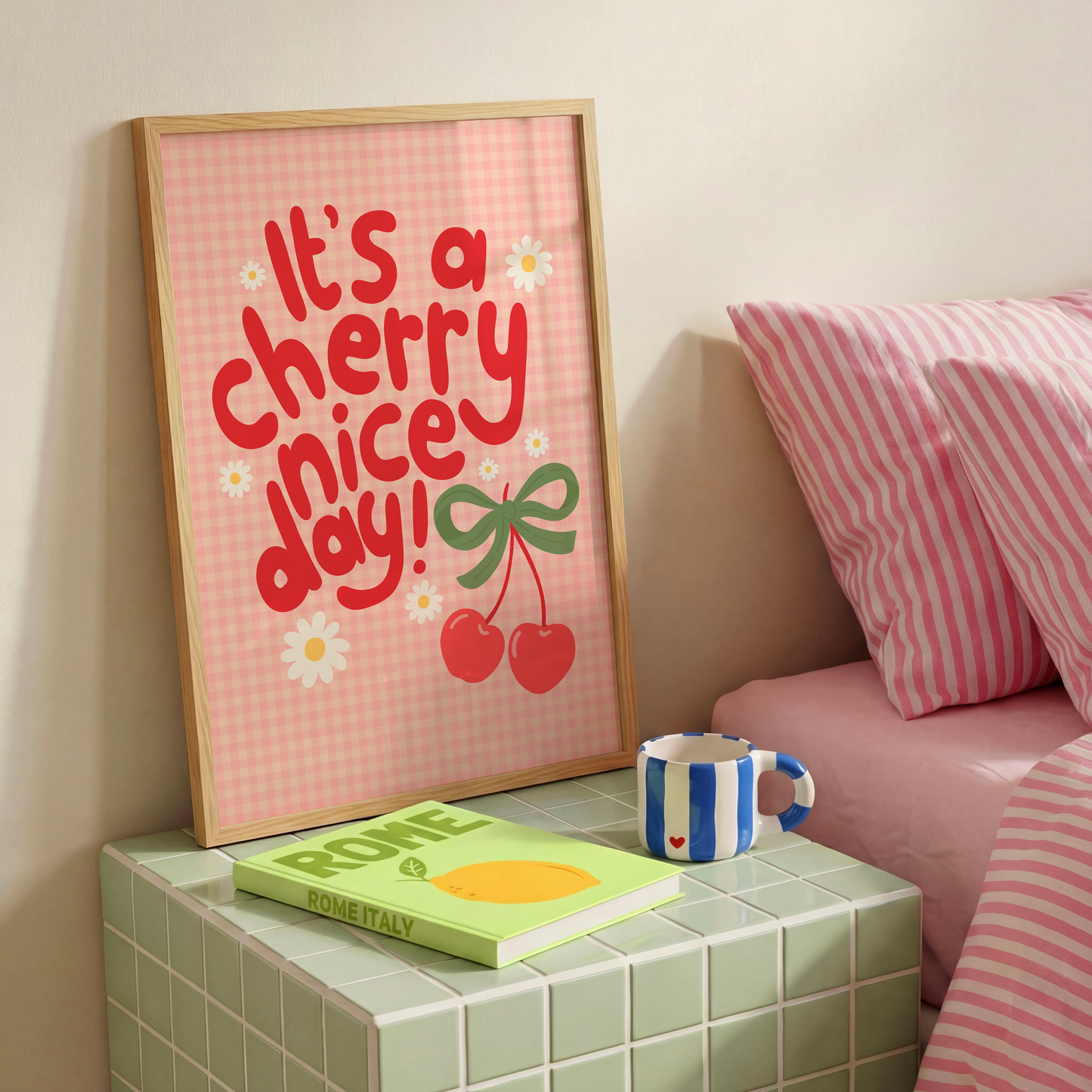 A gingham print, with hand lettered text that reads 'it's a cherry nice day!' in red. The text is surrounded by little daisy illustrations and a large pair of cherries tied together with a green bow.