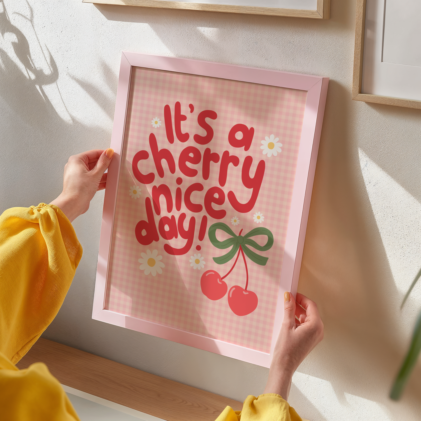 A gingham print, with hand lettered text that reads 'it's a cherry nice day!' in red. The text is surrounded by little daisy illustrations and a large pair of cherries tied together with a green bow.