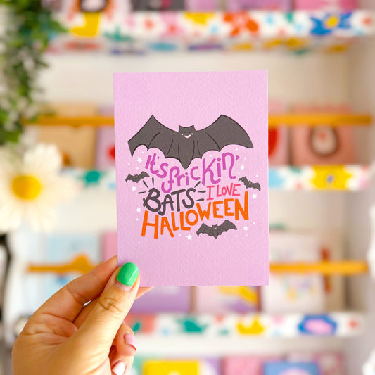 Pink card with Halloween text and bat design held by a hand in front of a colourful store display.