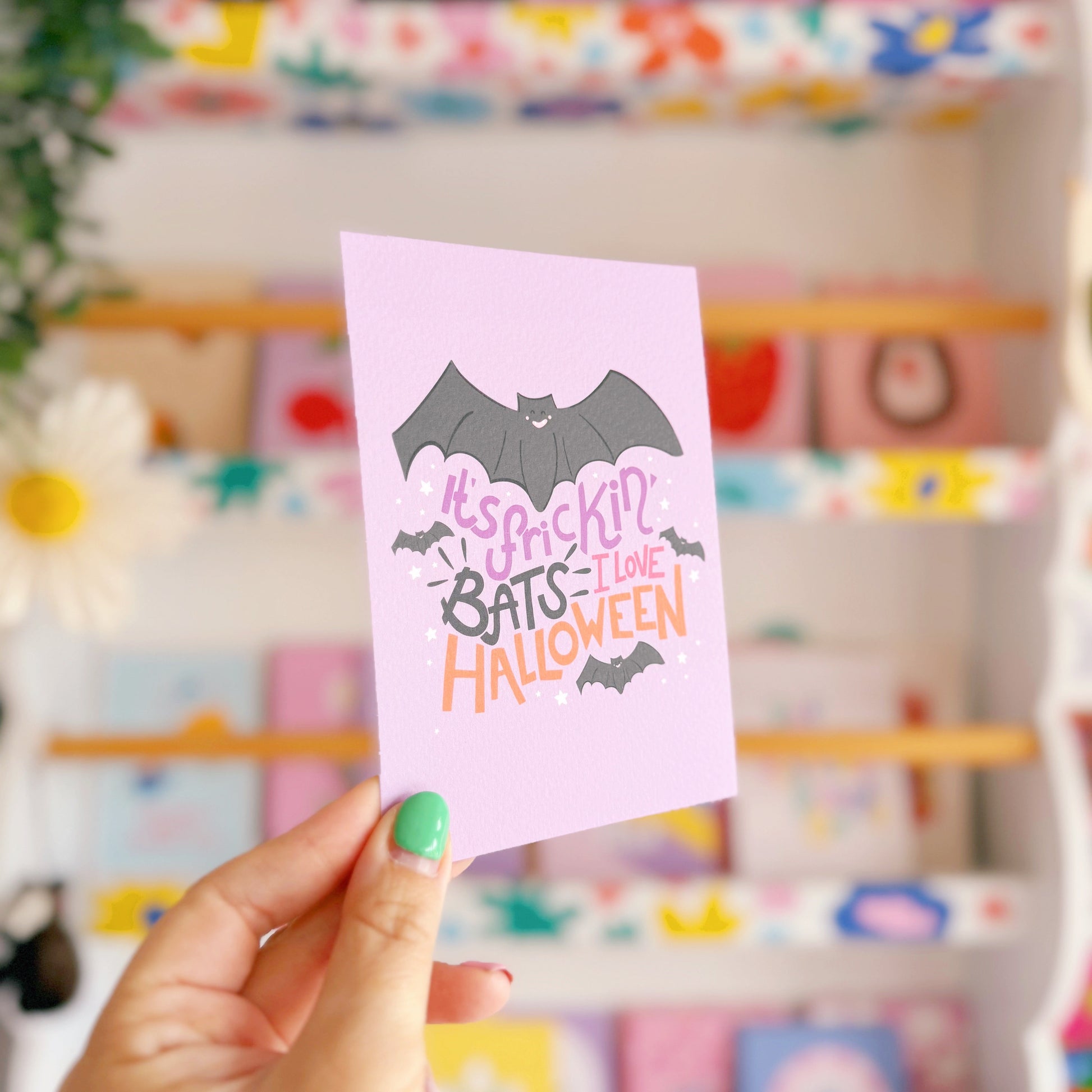 Hand holding a pink card with Halloween text and bat graphics in front of a colourful store display.
