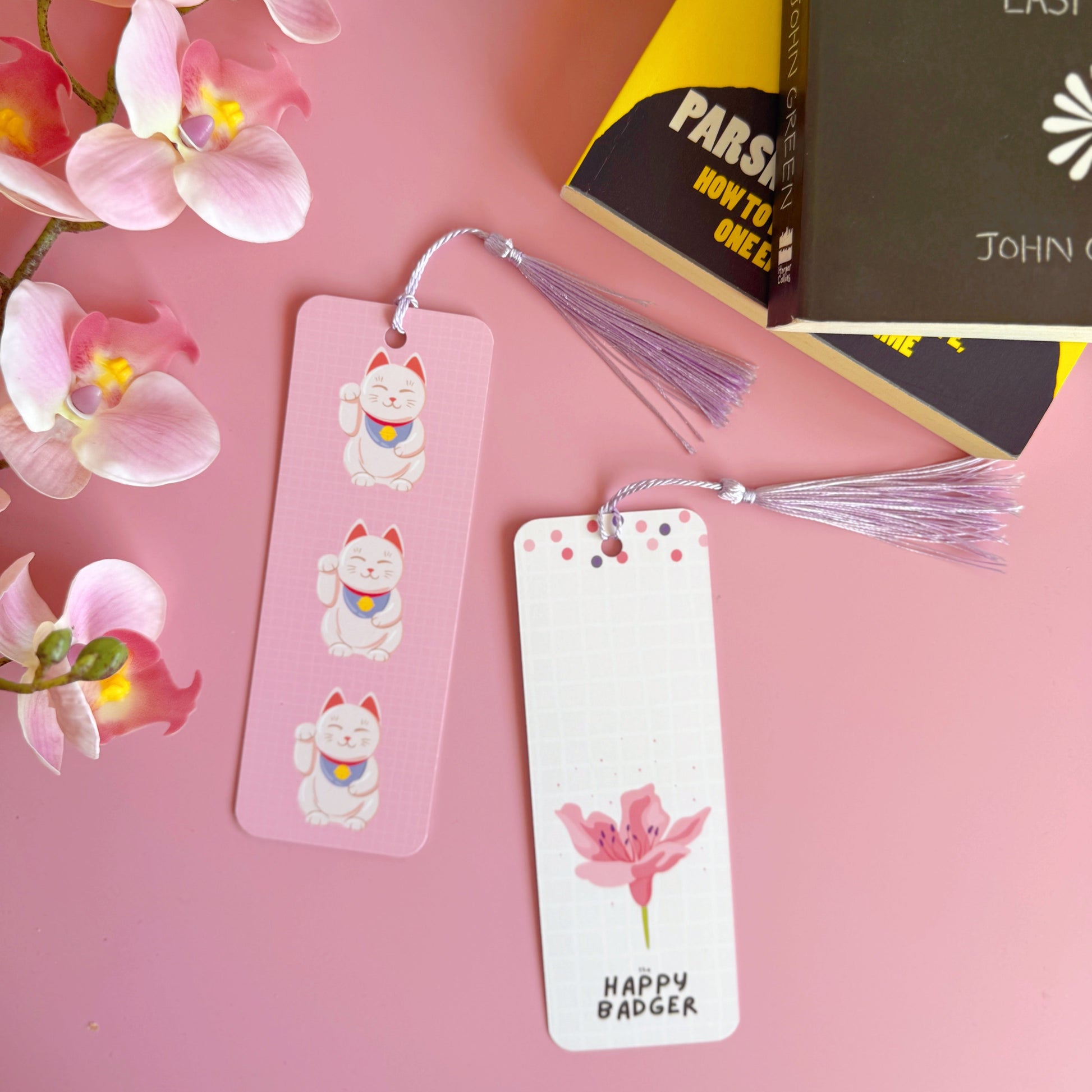 A pink checkered bookmark with three lucky cat illustrations on the front. Also shown is the back of the bookmark which is cream with a. pink cherry blossom and branding on. The bookmark has a lilac coordinating tassel.