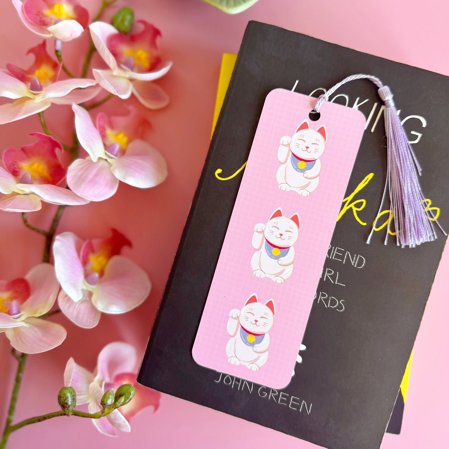 A pink checkered bookmark with three lucky cat illustrations on the front. The bookmark has a lilac coordinating tassel.