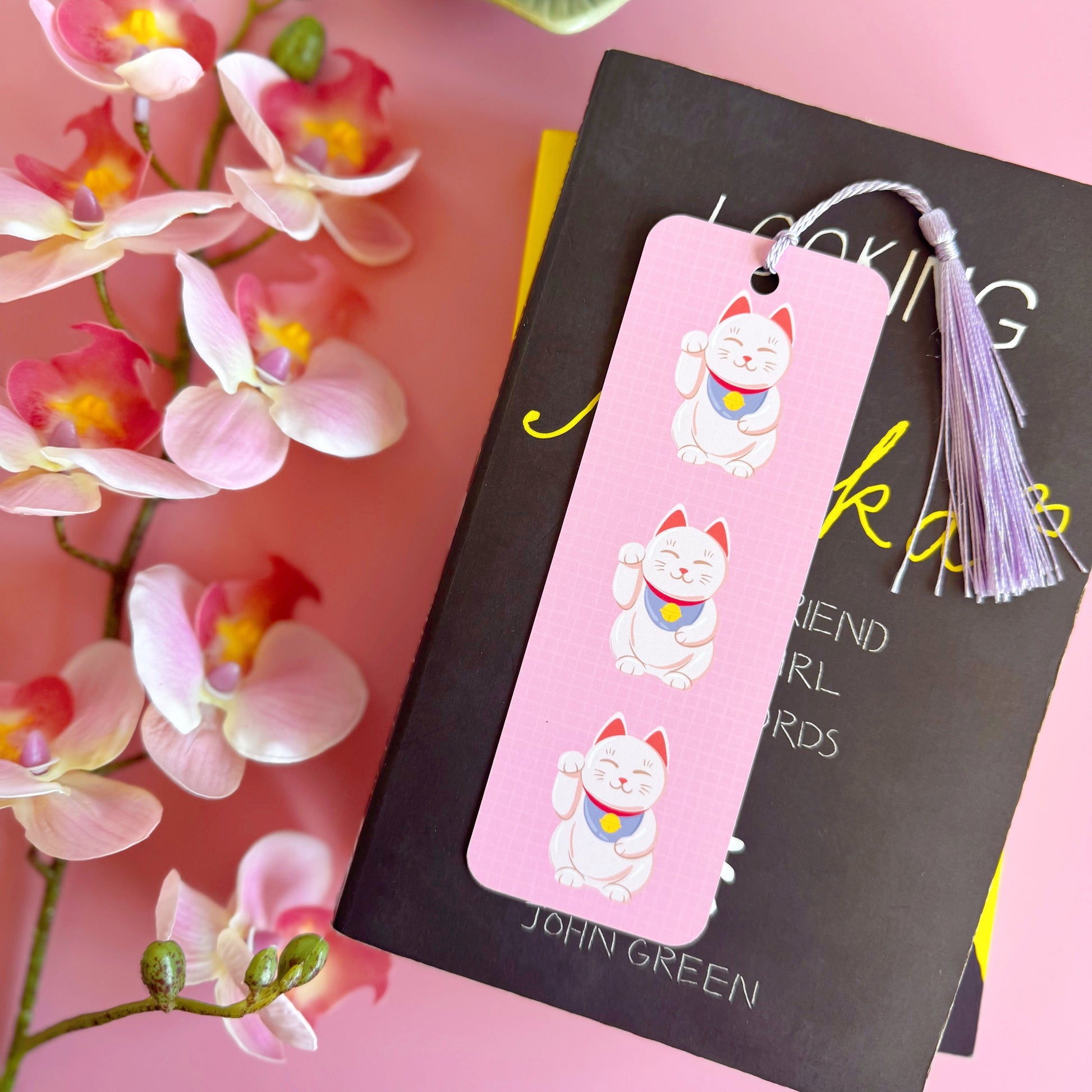 A pink checkered bookmark with three lucky cat illustrations on the front. The bookmark has a lilac coordinating tassel.