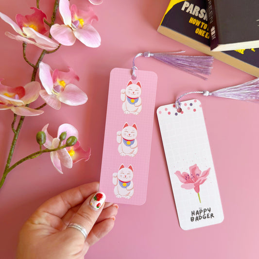 A pink checkered bookmark with three lucky cat illustrations on the front. Also shown is the back of the bookmark which is cream with a. pink cherry blossom and branding on. The bookmark has a lilac coordinating tassel.