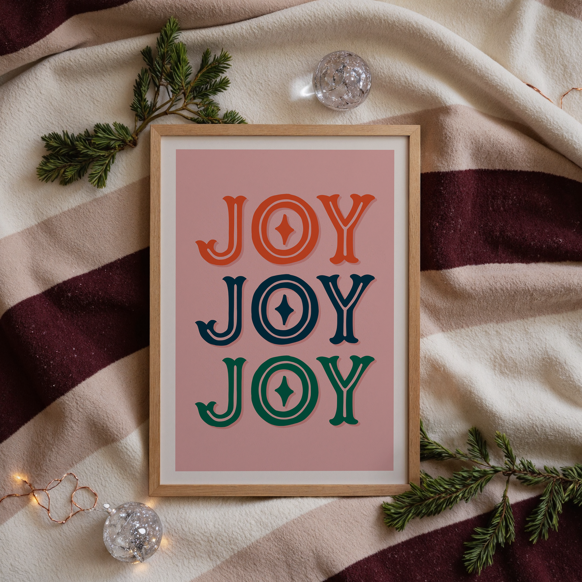 A print with a warm pink background and the word 'joy' written, in an ornate text, three times - one in red, one in navy and one in green.