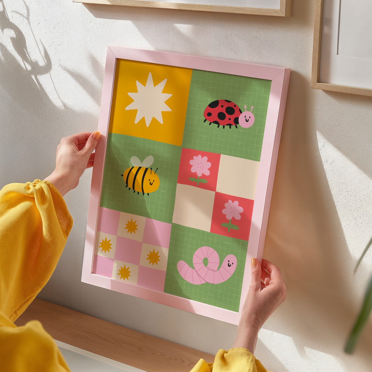 A framed children's art print. The illustration features colourful cartoon motifs including a ladybug, bee, worm, flowers, and suns on green, pink, yellow, and cream squares.