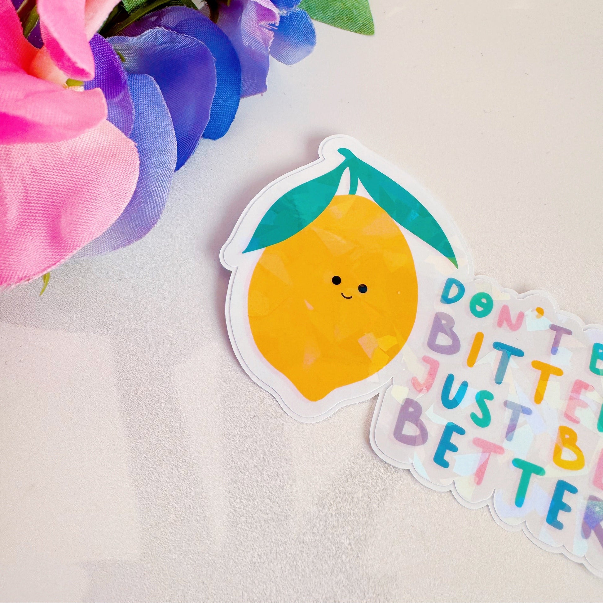 A holographic sticker, of a smiling lemon and the quote don't be bitter just be better. The sticker has an easy peel lip.