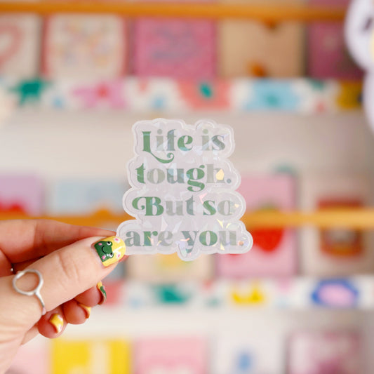 A green holographic finish sticker, with green text saying 'life is tough but so are you'.