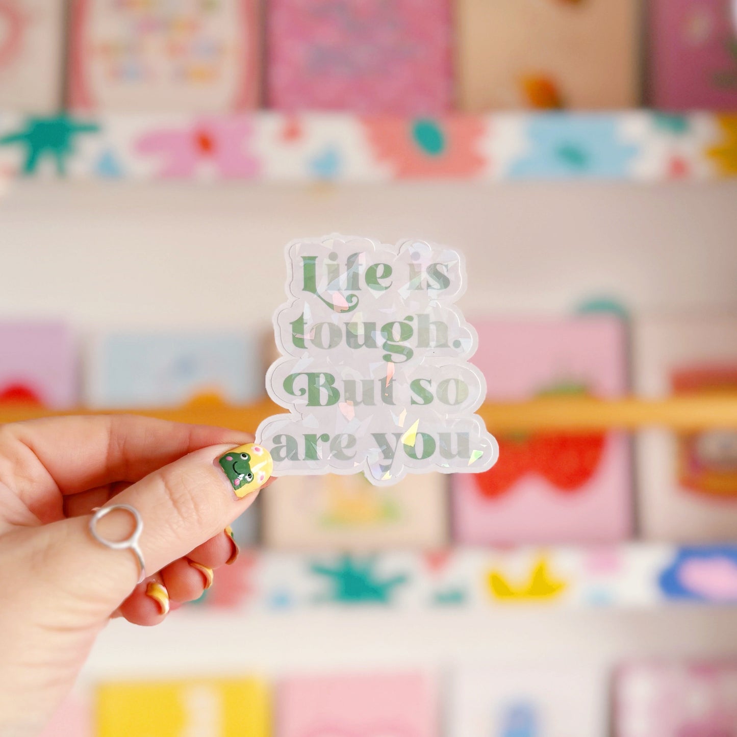 A green holographic finish sticker, with green text saying 'life is tough but so are you'.