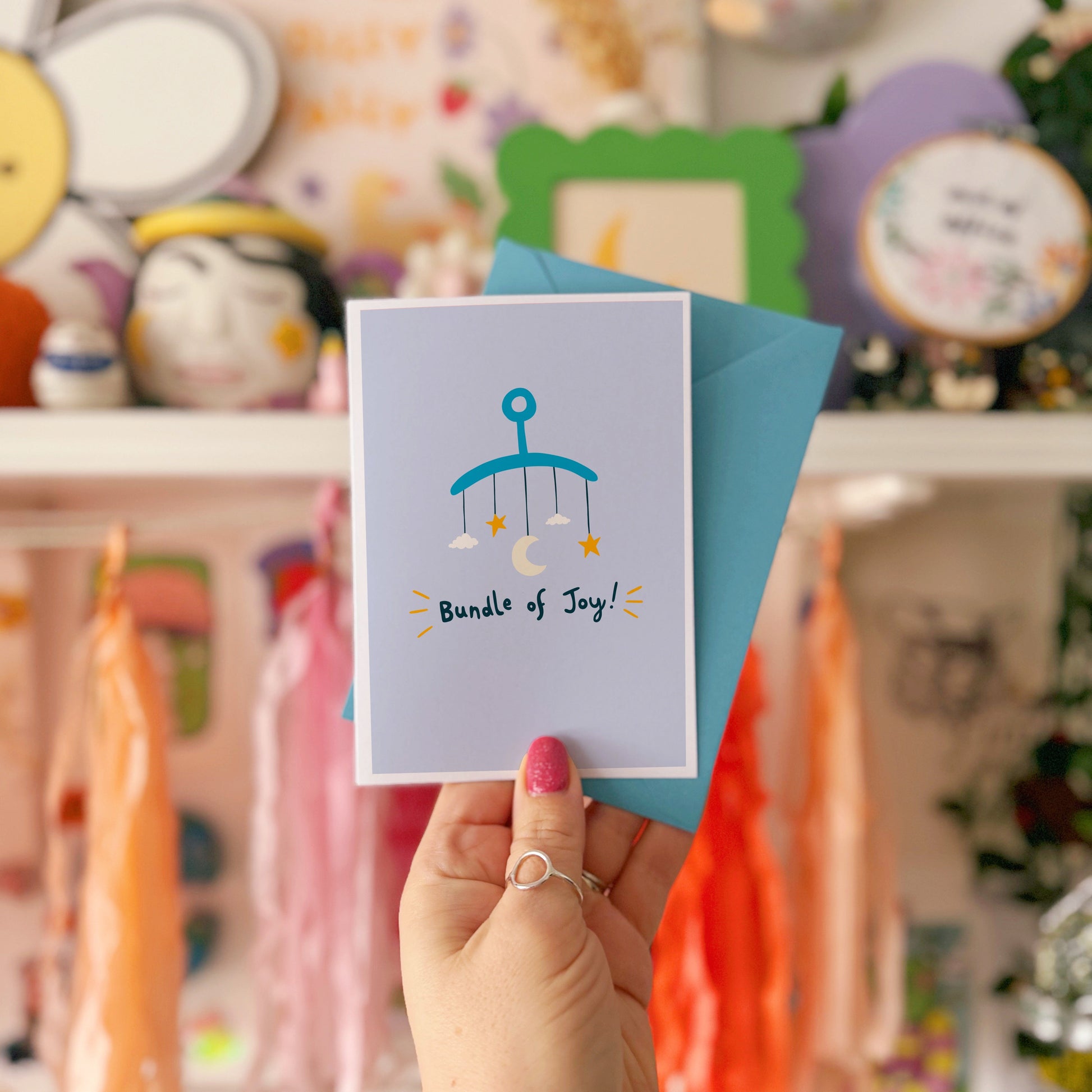 A blue greeting card with an illustration of a mobile. Underneath the illustration is hand written text that reads 'bundle of joy!'.