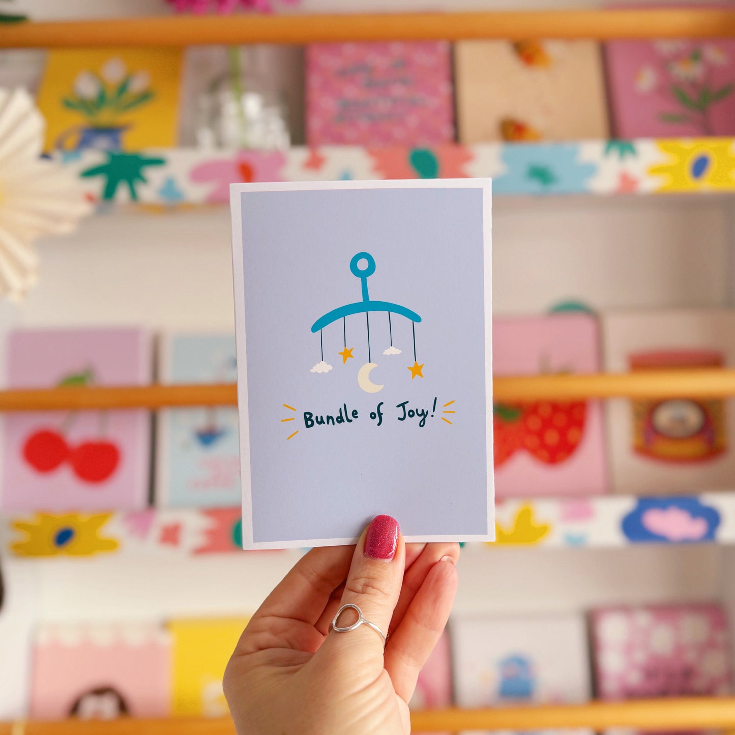A blue greeting card with an illustration of a mobile. Underneath the illustration is hand written text that reads 'bundle of joy!'.