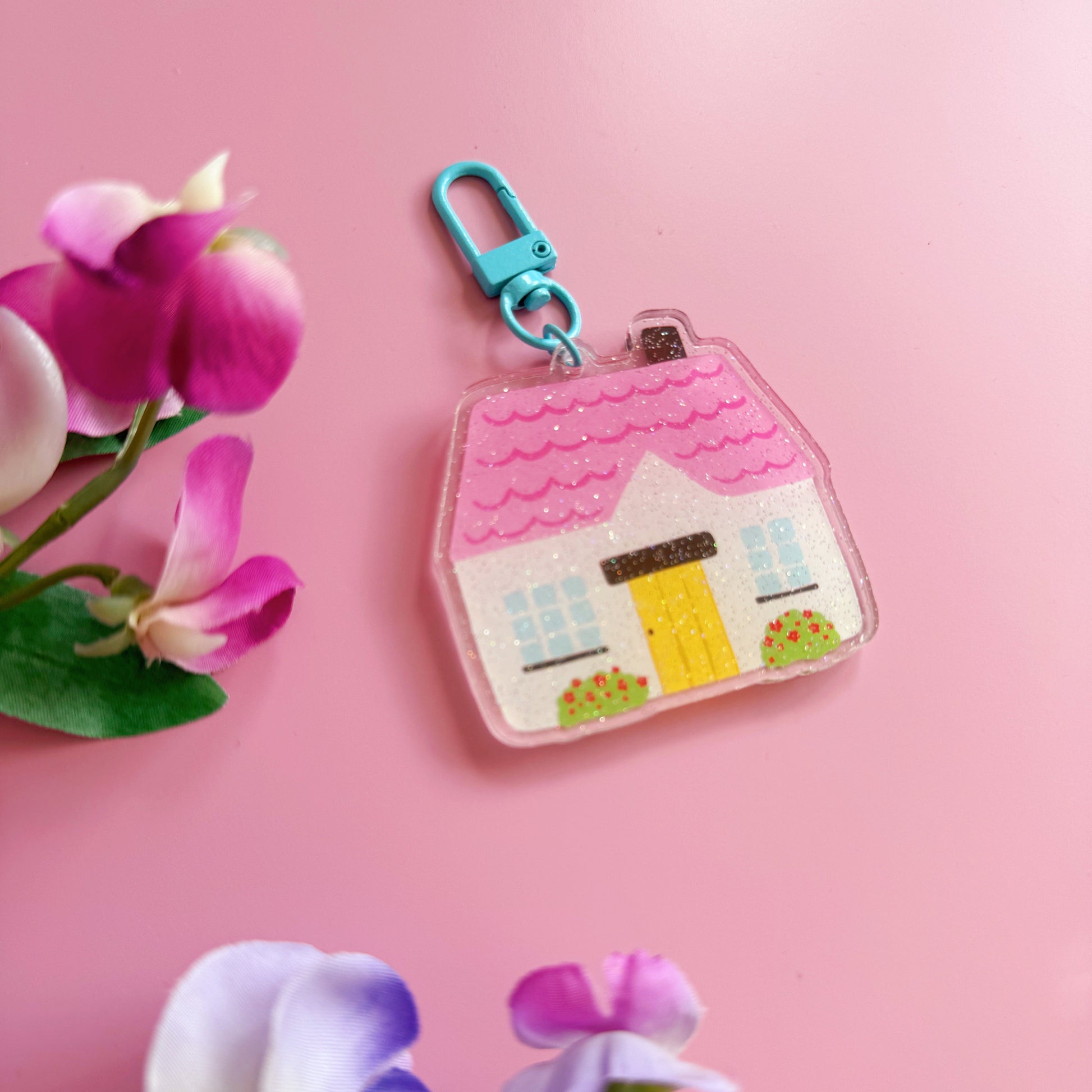 House-shaped keychain with a pink roof and yellow door on a pink background with flowers.