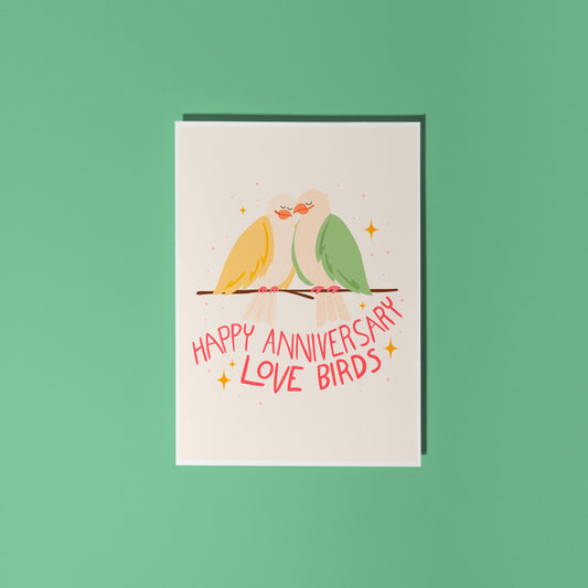 An illustrated anniversary card with two birds sitting on a branch, looking loved up. The hand written lettering underneath them reads ;happy anniversary love birds'.