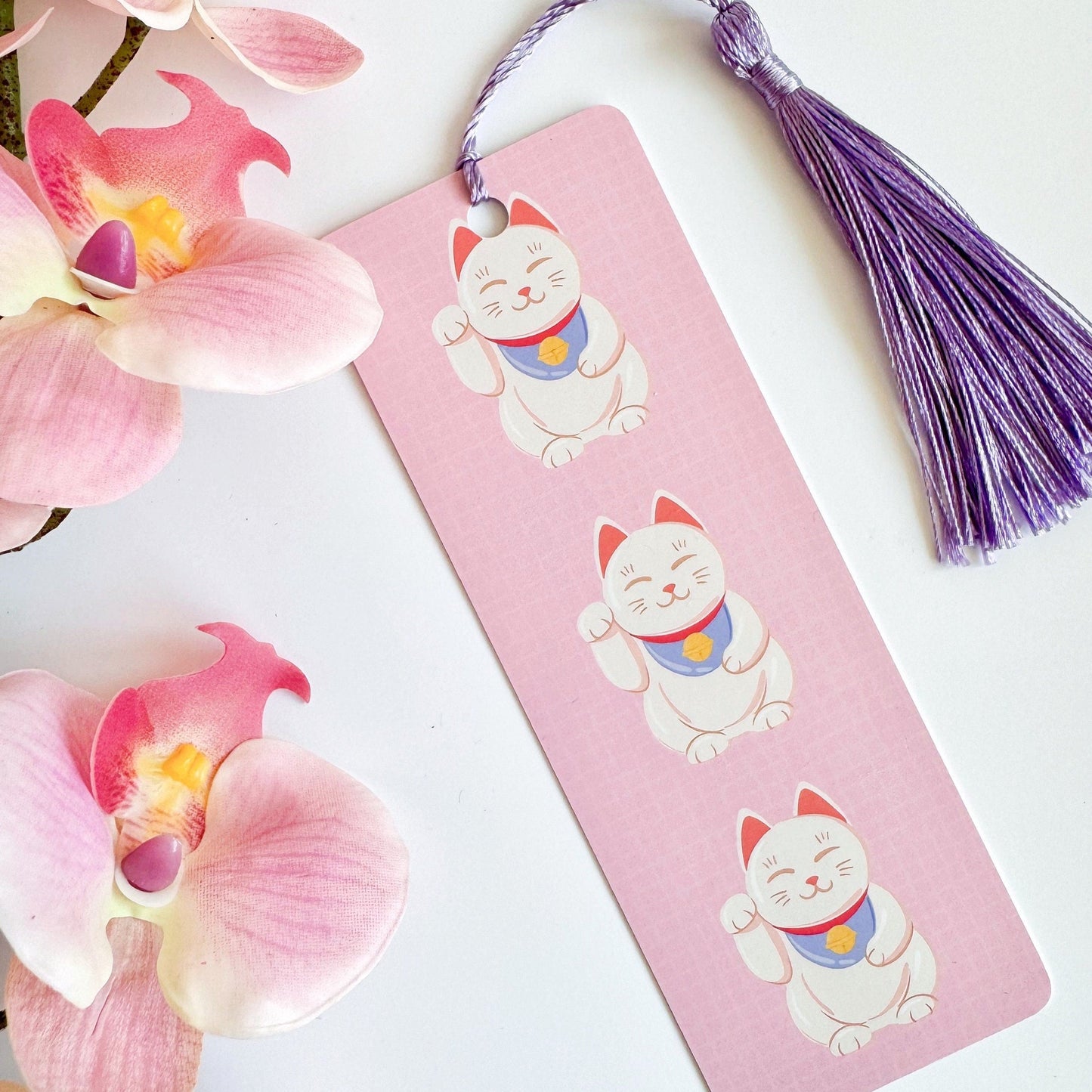 A picture of a pink bookmark, with a coordinating purple tassel, with three illustrations of Japanese lucky cats on the front.