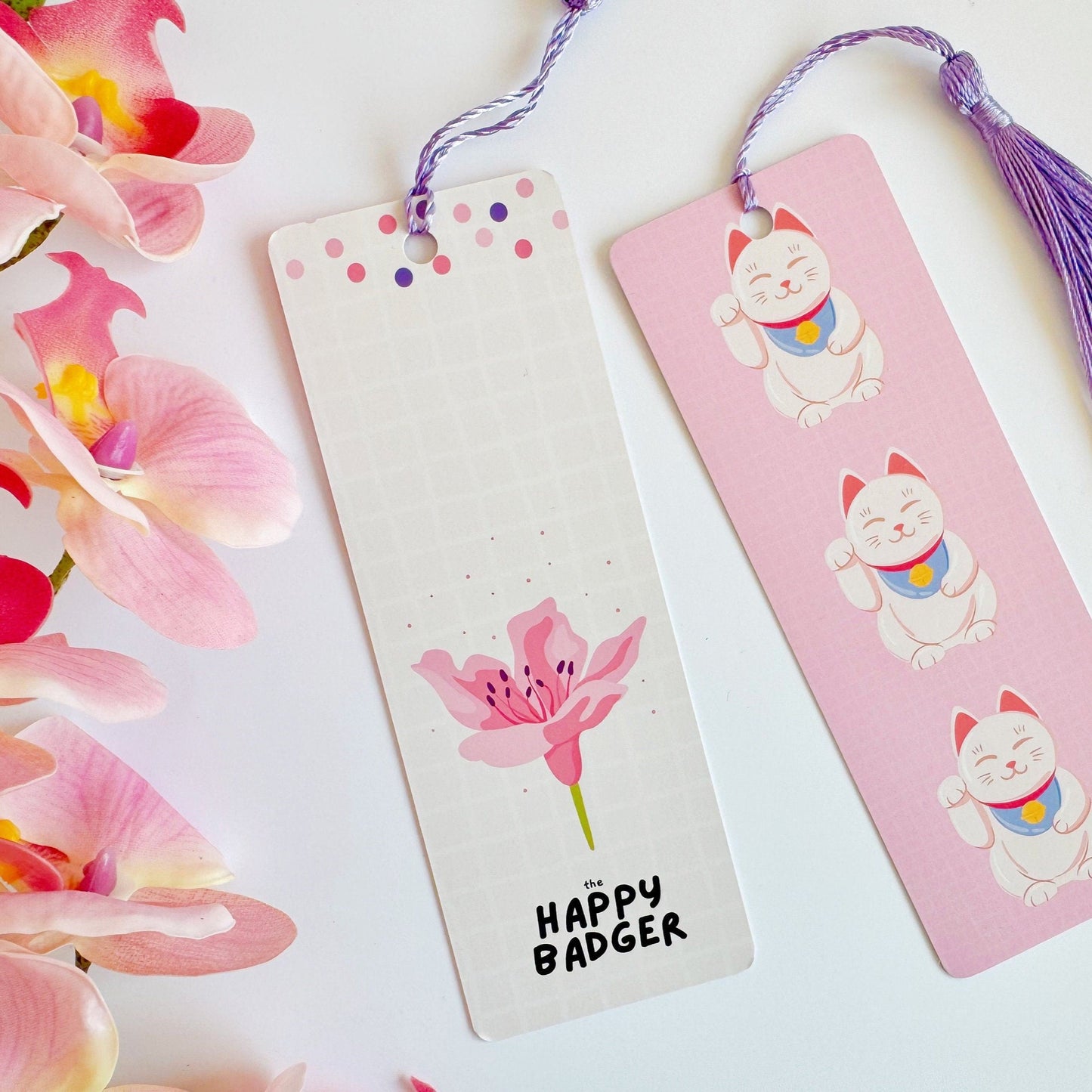 A picture of a pink bookmark, with a coordinating purple tassel, with three illustrations of Japanese lucky cats on the front. Also shown is the rear side, which is cream with a cherry blossom illustration and the brand logo to the bottom.