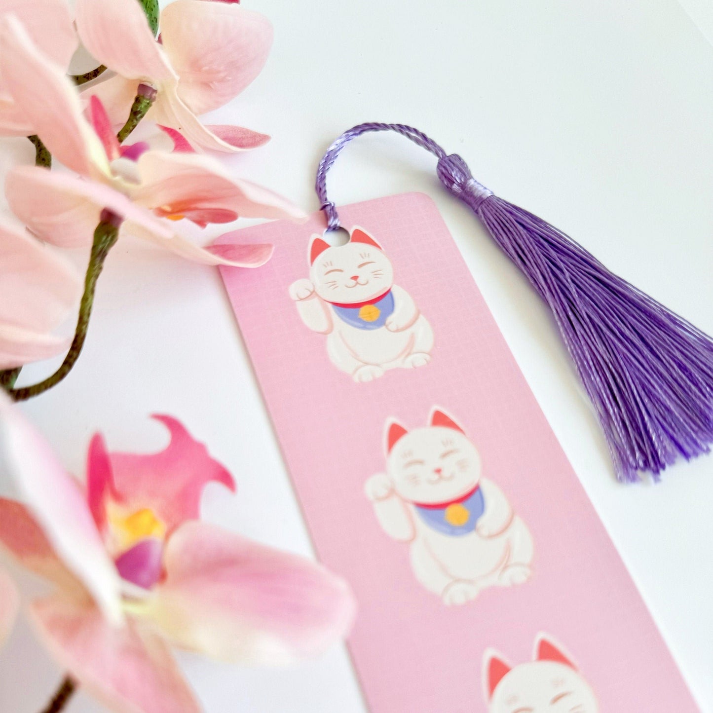 A picture of a pink bookmark, with a coordinating purple tassel, with three illustrations of Japanese lucky cats on the front.