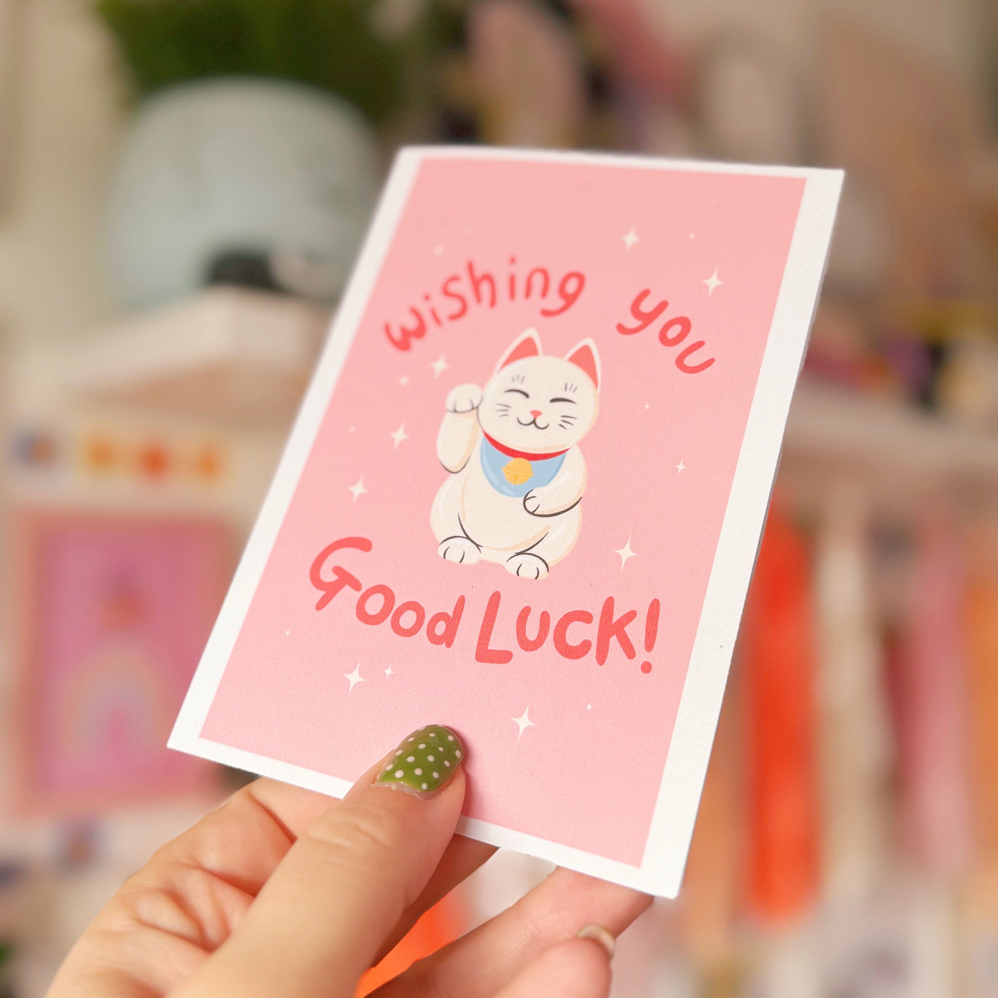 Hand holding a pink card with a cat illustration and 'Wishing you Good Luck!' text.